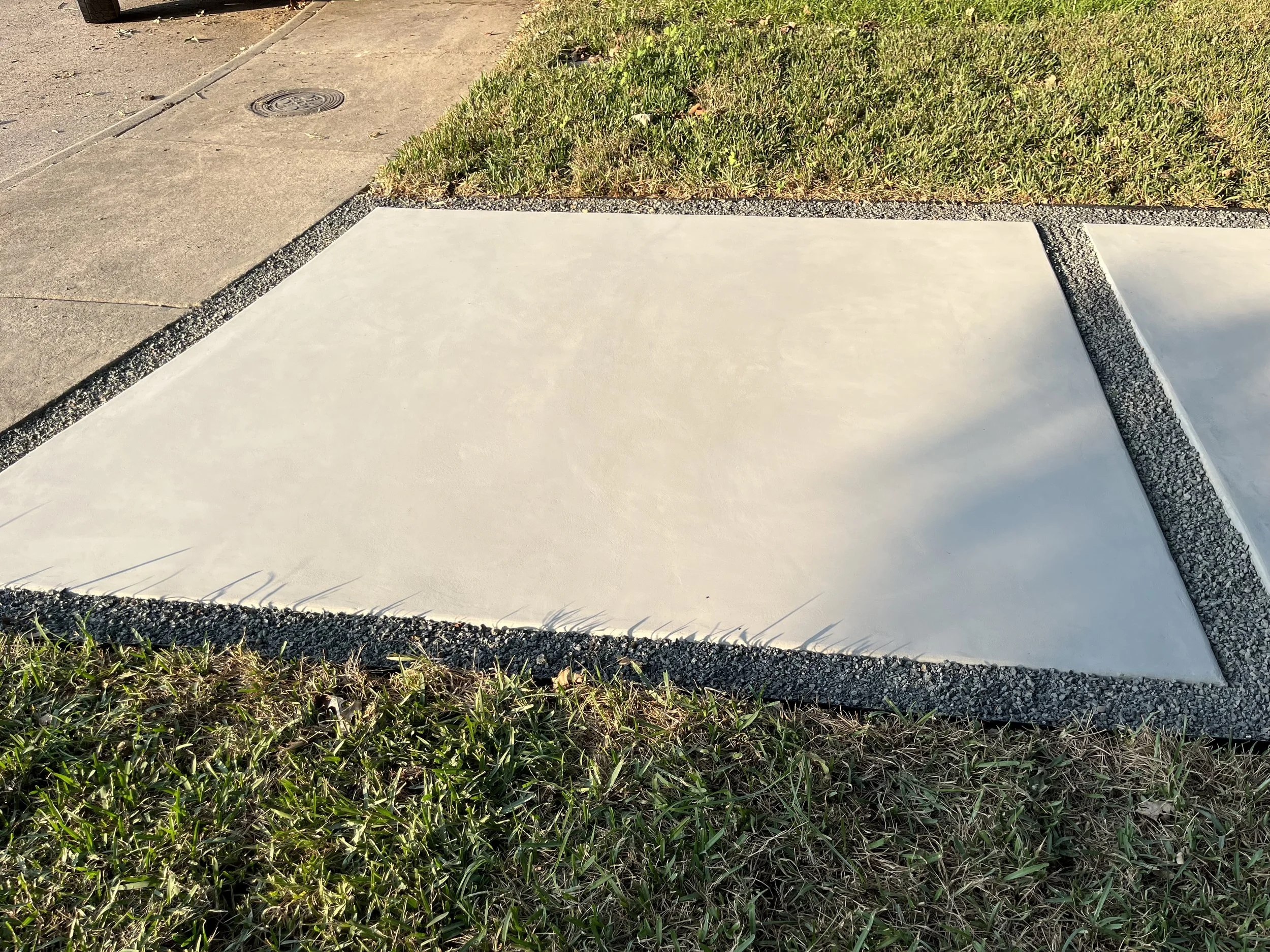Concrete sidewalk patch surrounded by grass and adjacent to a paved sidewalk with a drain cover.
