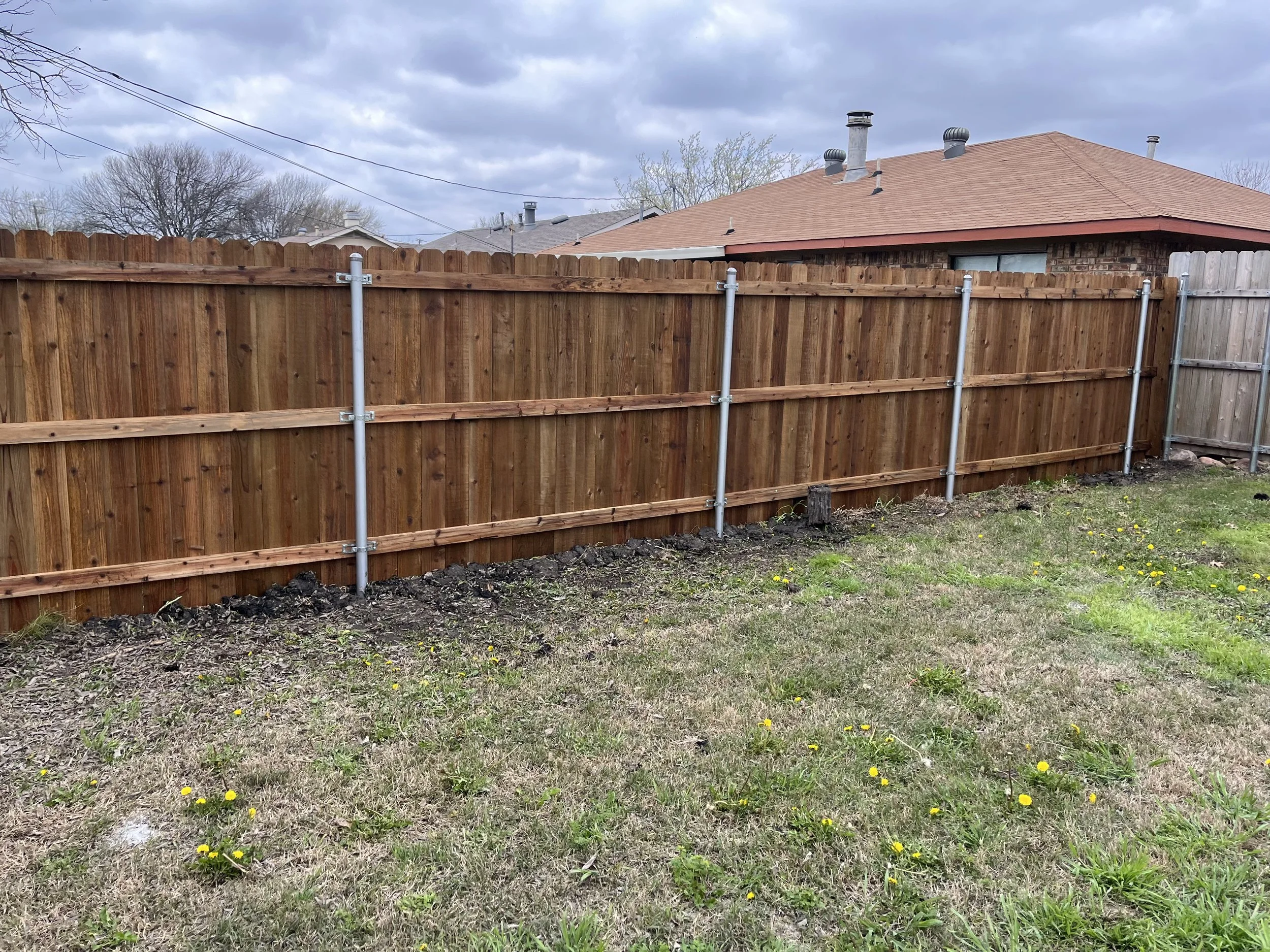 A newly installed wooden privacy fence with metal posts in a backyard with patchy grass and small yellow flowers, under cloudy skies.