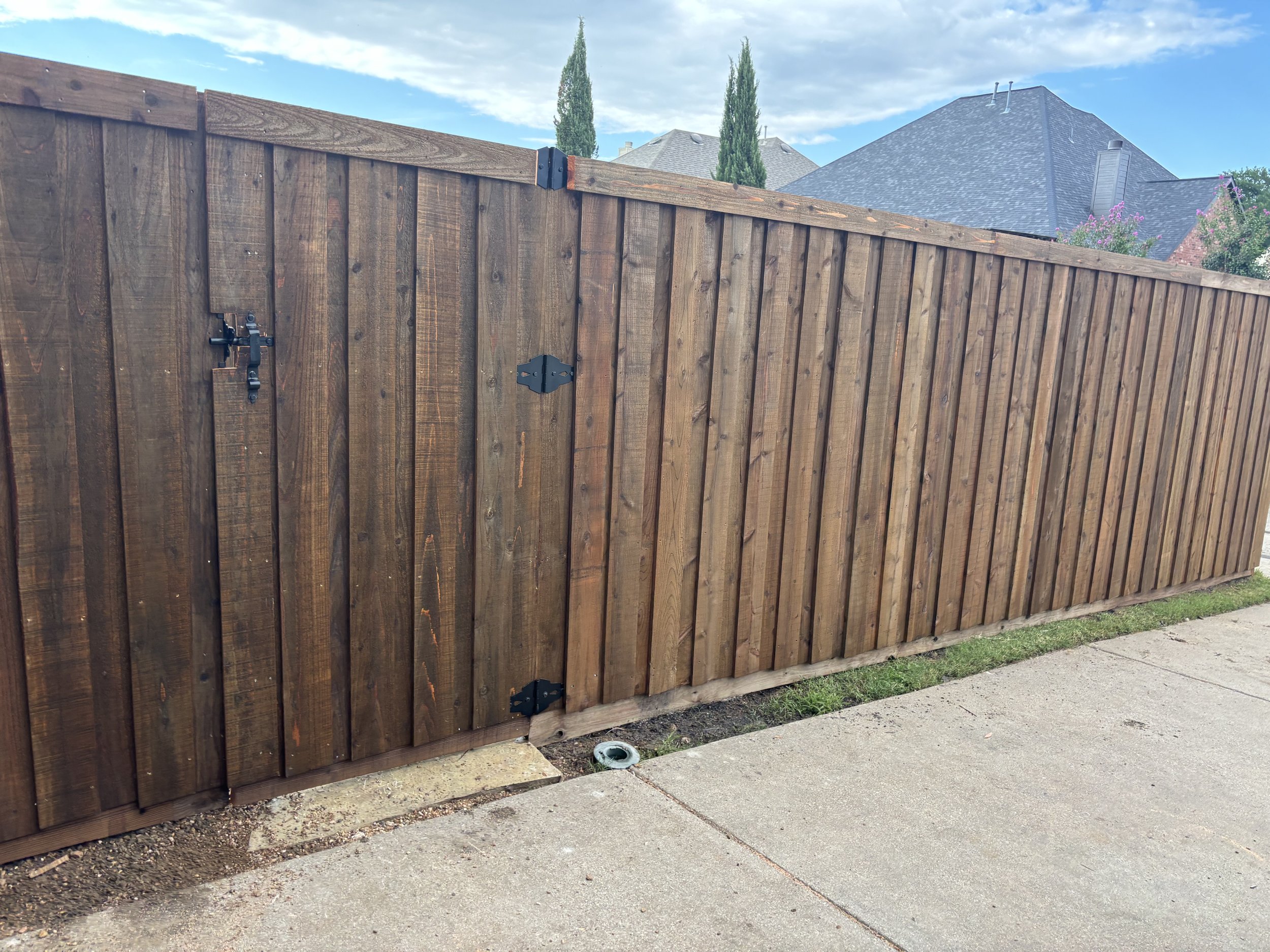 Wooden privacy fence with black hardware, adjacent to a sidewalk, with house rooftops and blue sky in the background.