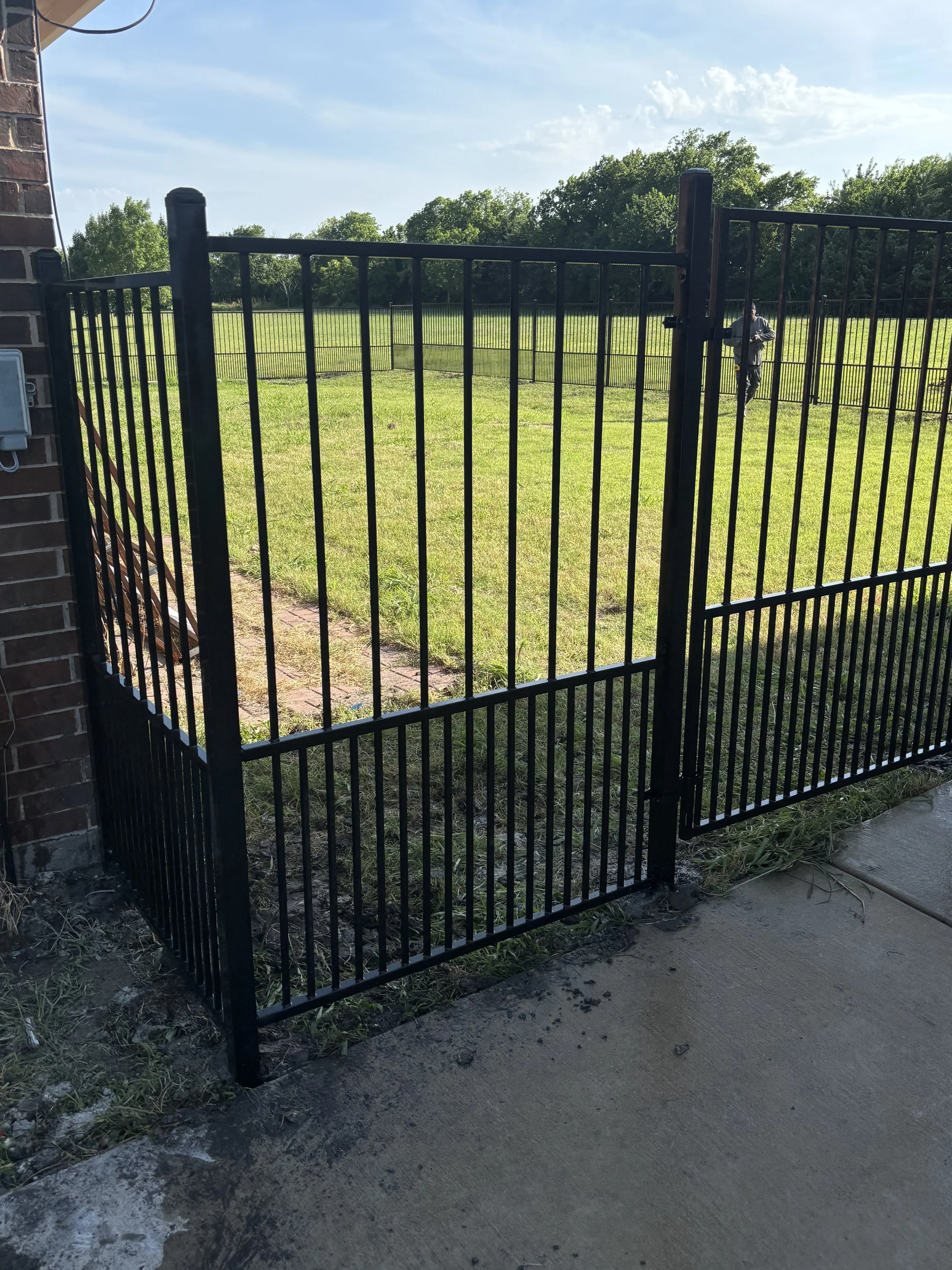 Black metal gate attached to a brick wall opening to a grass field with trees in the background. A person is visible in the distance on the right side of the image.