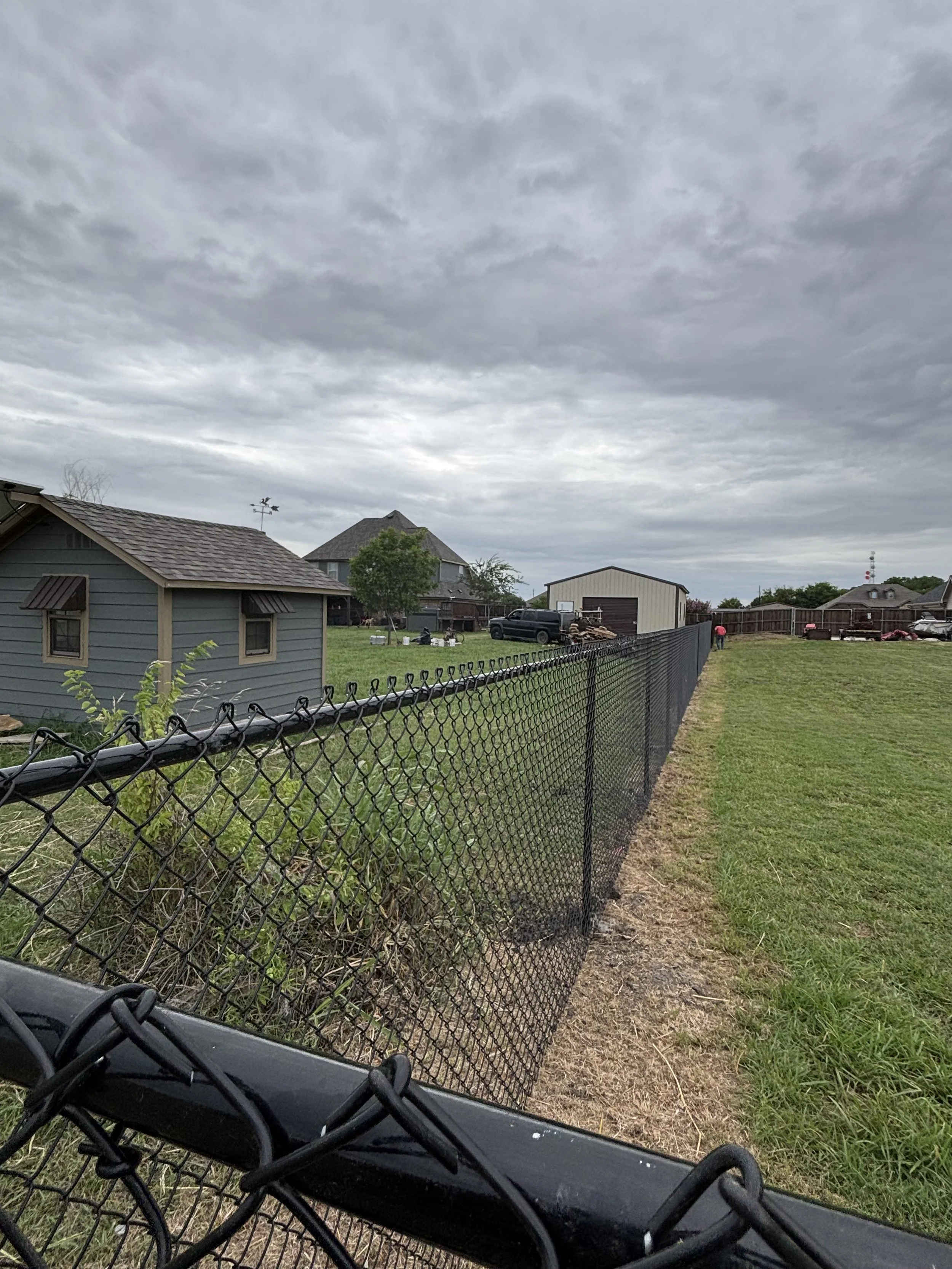 A residential backyard with a black chain-link fence, a small gray shed, a lawn, and houses in the distance under a cloudy sky.