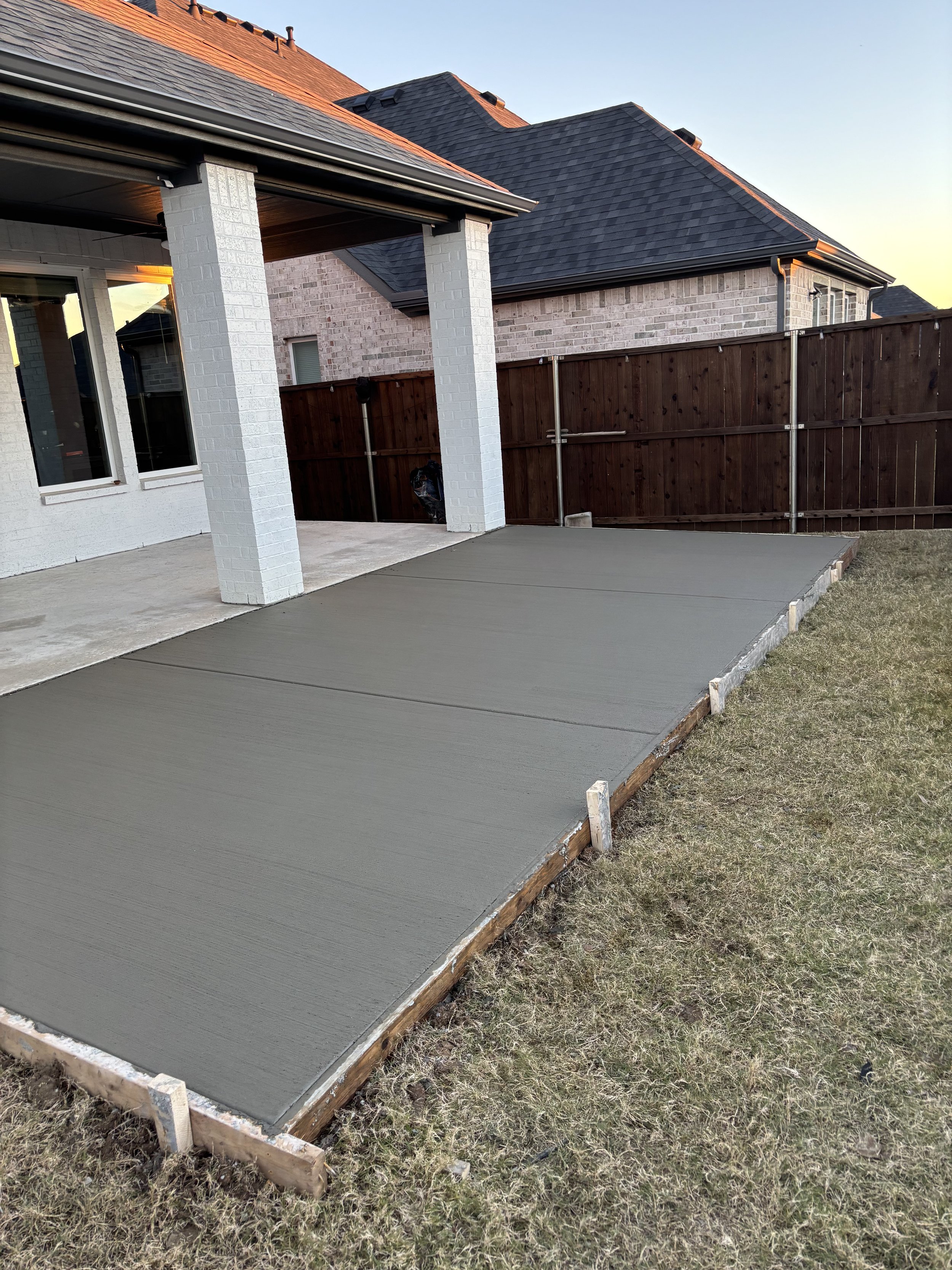 Freshly poured concrete patio in backyard with a wooden frame, adjacent to a lawn, next to a house with brick exterior and large windows, enclosed by a wooden fence.
