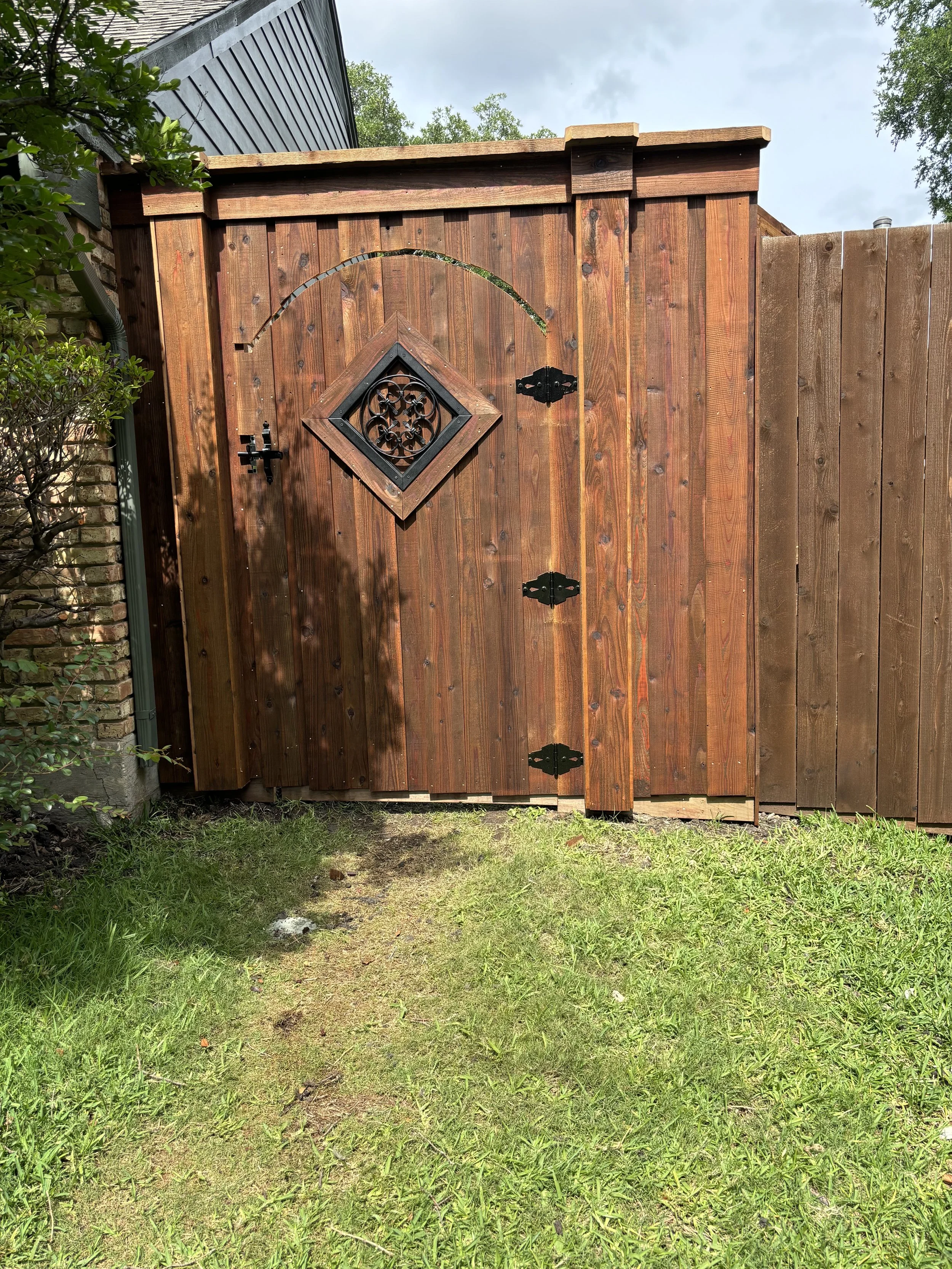 A wooden backyard gate with a decorative diamond-shaped window and black hinges, surrounded by green grass and shrubbery.