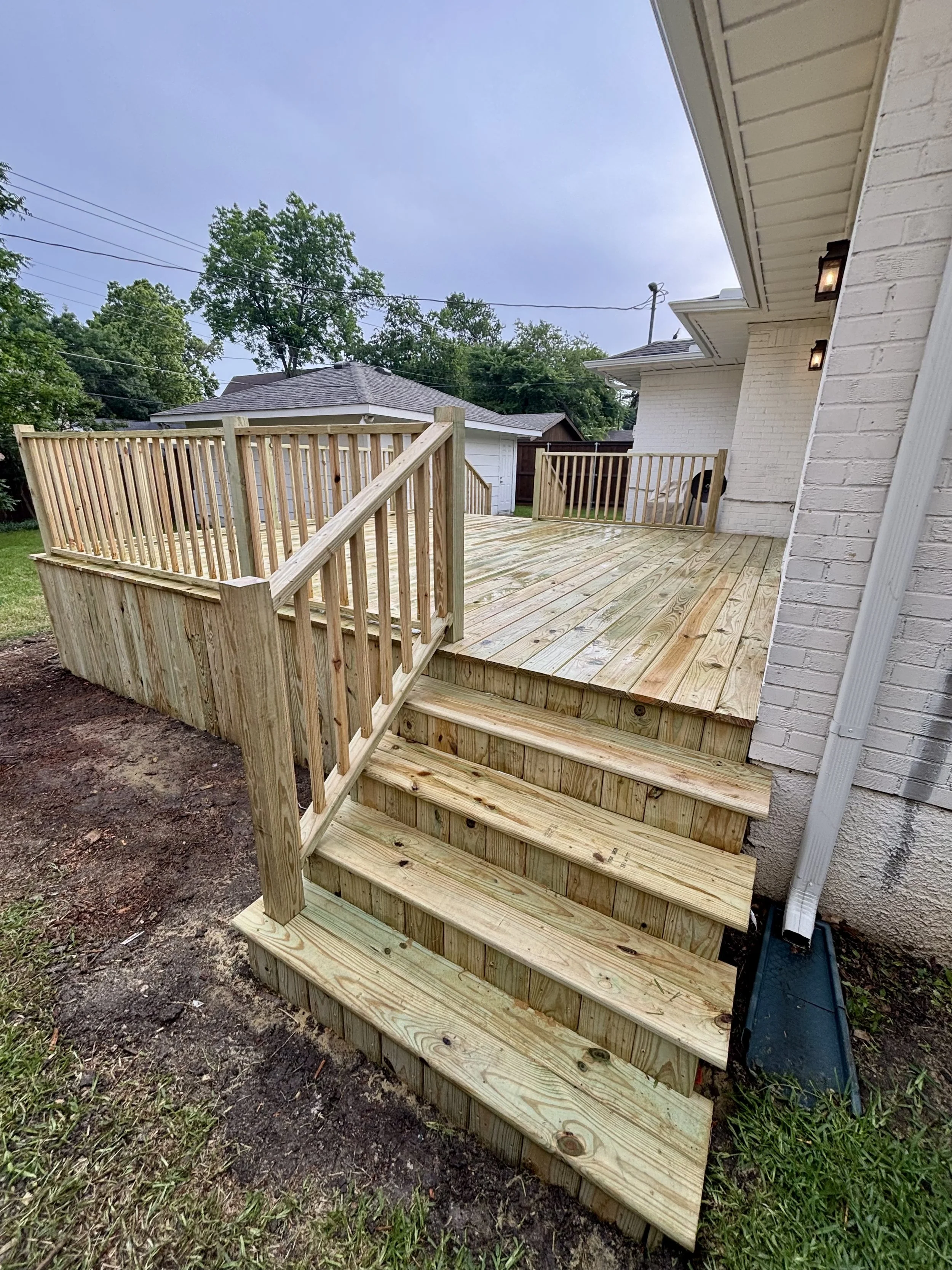 New wooden deck with stairs and railing attached to a white brick house, outdoor lighting, backyard with trees and neighboring houses in the background.