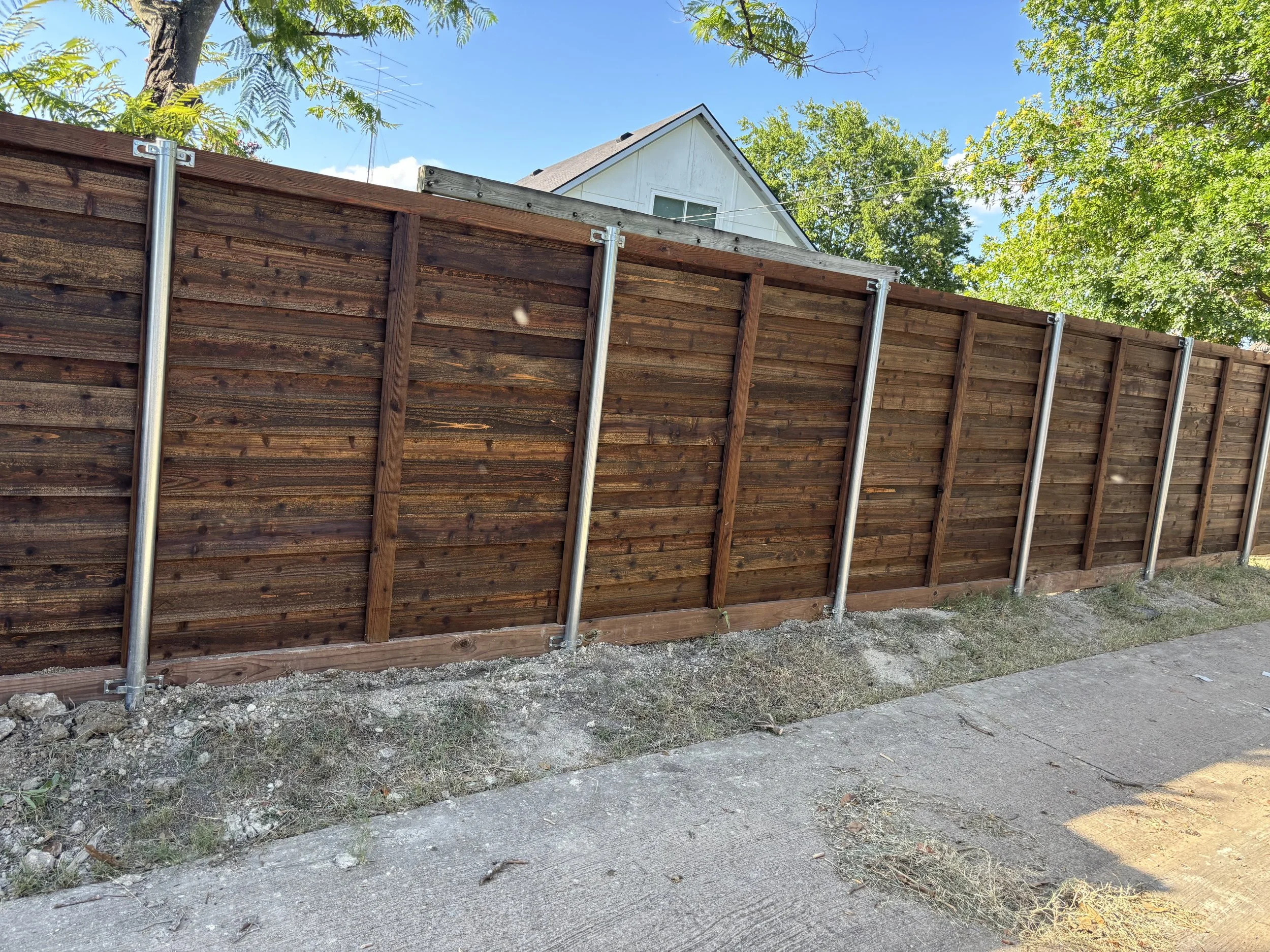 Wooden privacy fence with metal support stakes along sidewalk in front of a residential house with trees and blue sky in background.