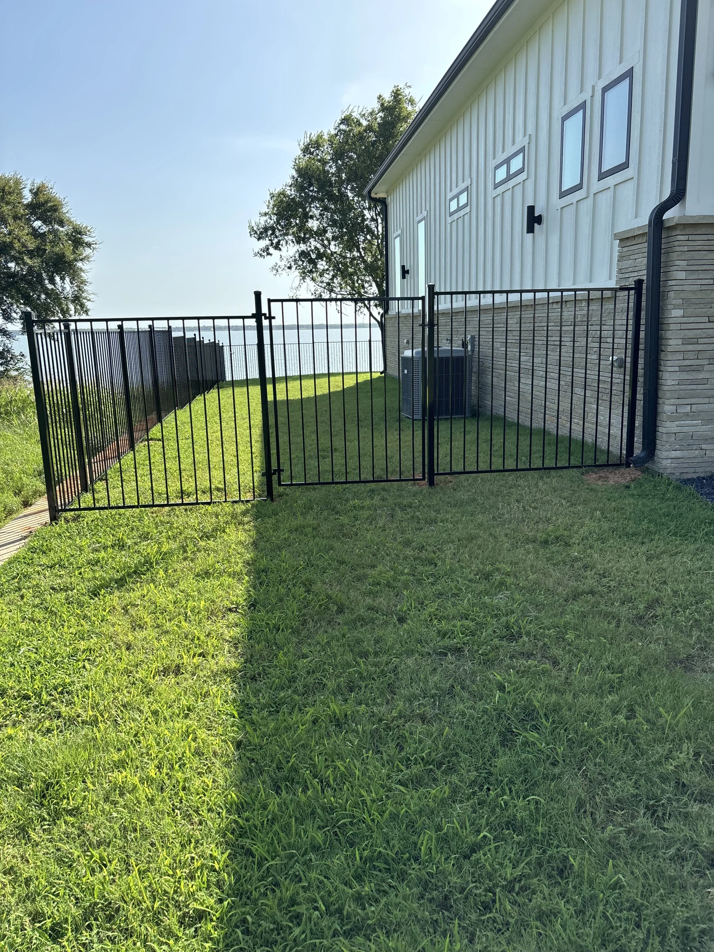 A black metal gate with a latch, part of a fence, on a grassy lawn beside a house with white siding and brick foundation. The house has multiple small windows, a black gutter, and an air conditioning unit outside. In the background, there is a body o