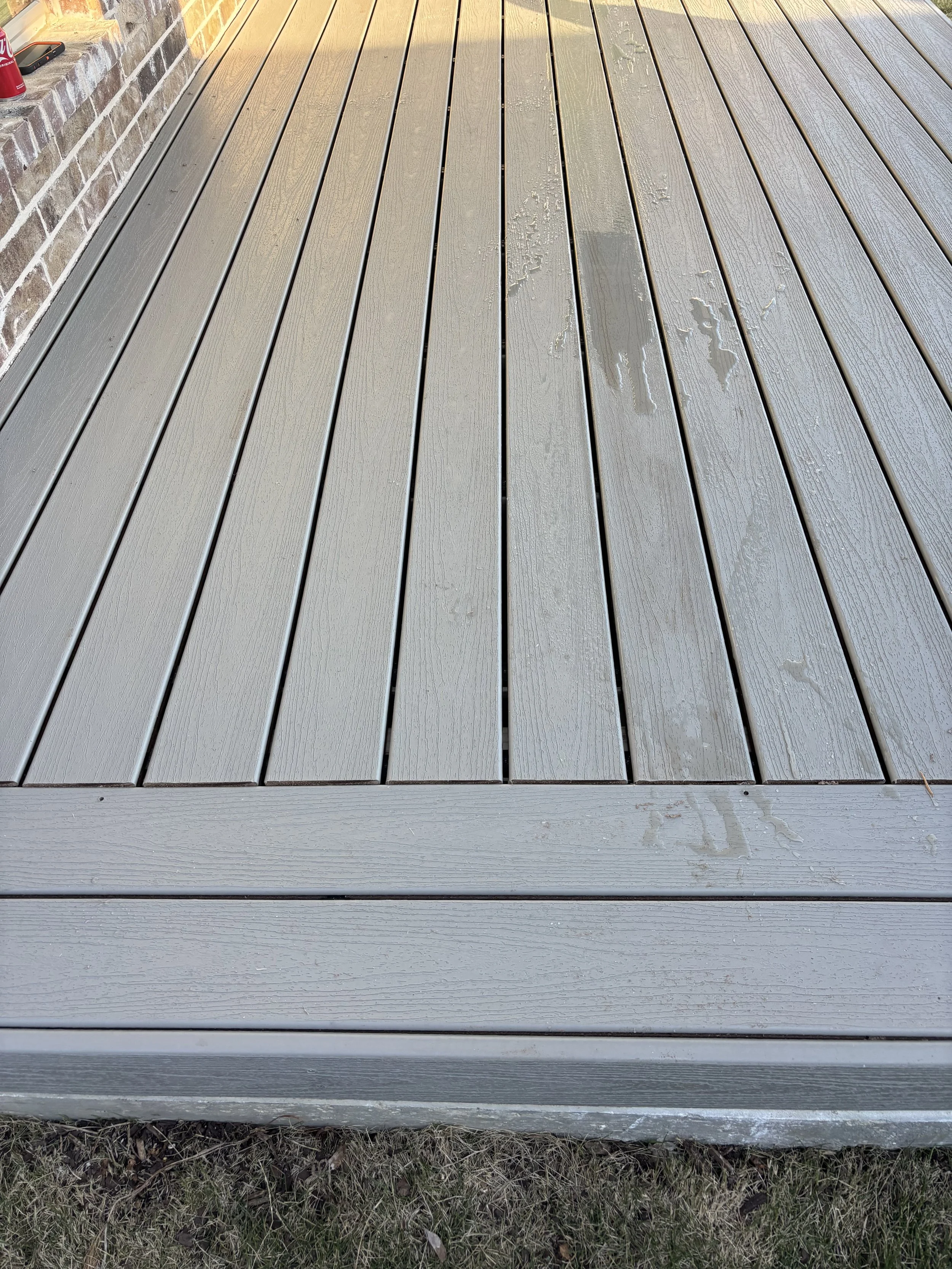 Close-up of wet gray wooden deck with water spots, adjacent to a brick wall on the left, and some grass at the bottom.