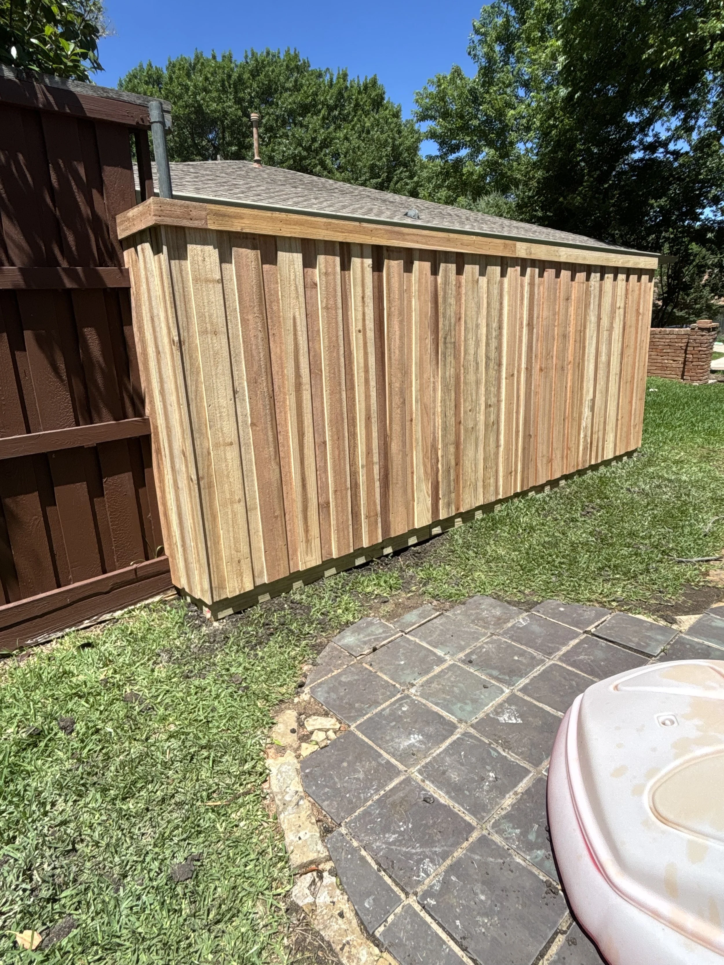 A wooden fence with vertical planks on a backyard, next to a brown fence, with green grass and a paved stone area in the foreground, and Tall green trees with clear blue sky in the background.