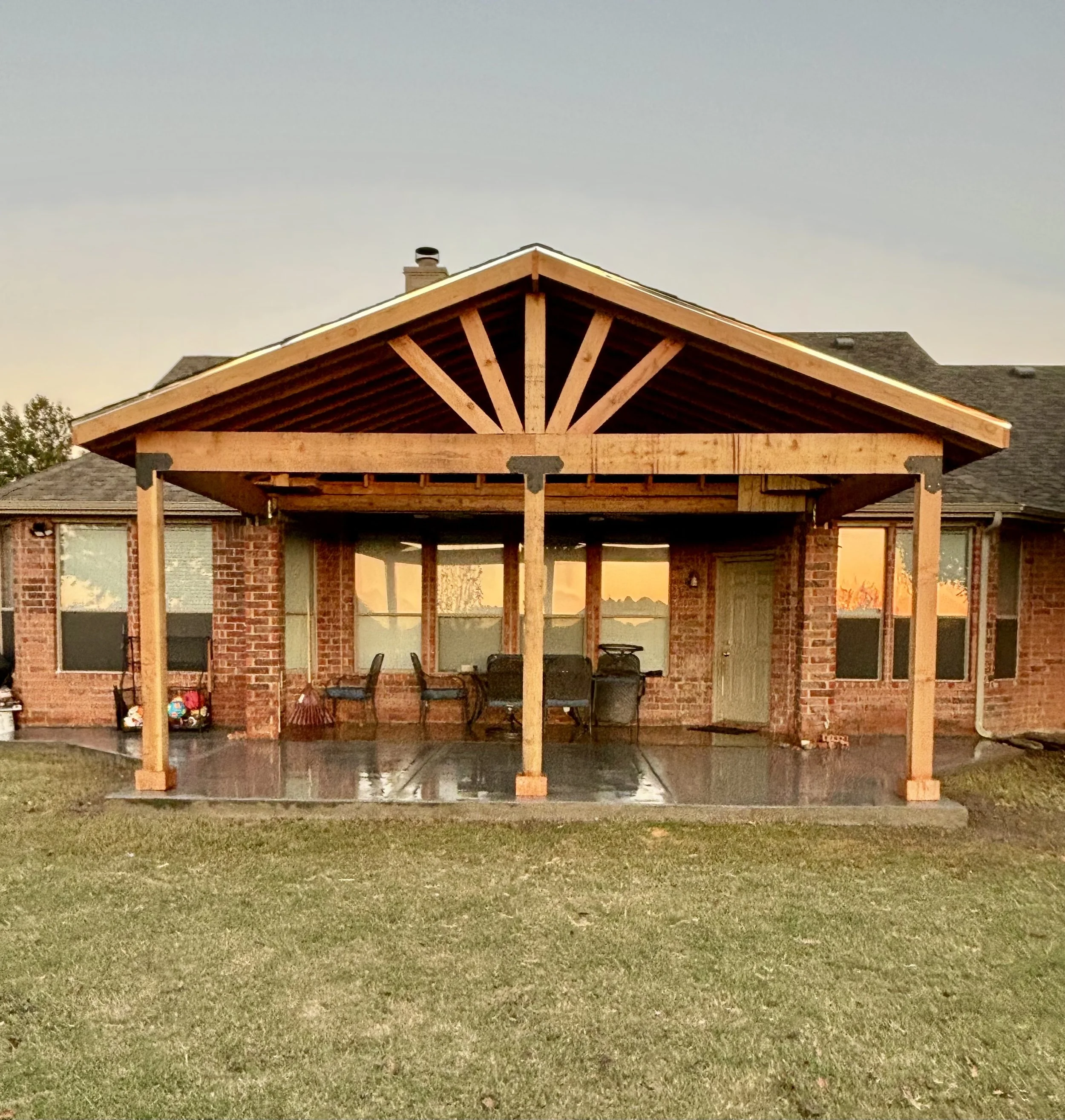 Back porch of a brick house with a new wooden pergola structure, outdoor table and chairs, and a grassy lawn.