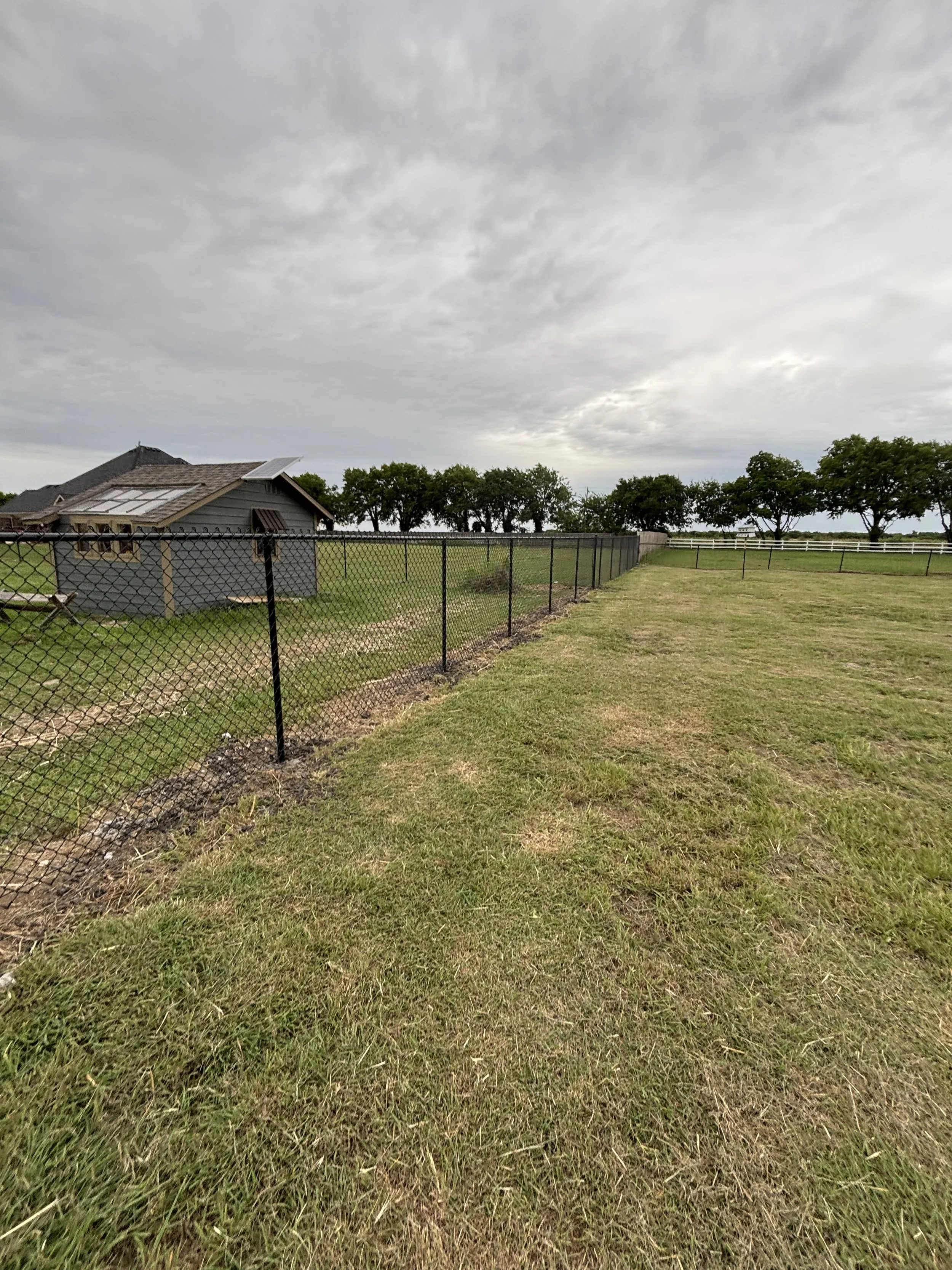 A fenced grassy yard with a small gray house on the left and trees in the background under a cloudy sky.