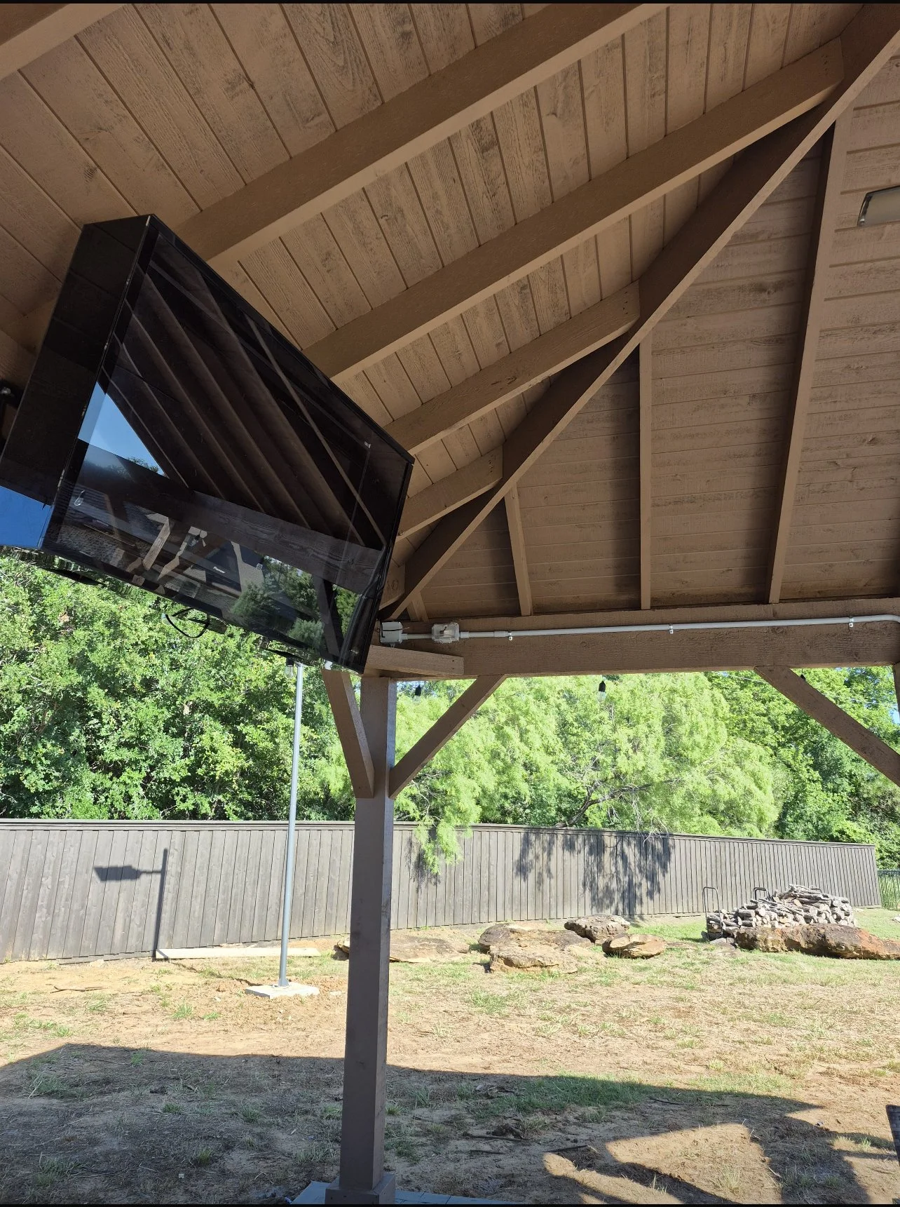 View of an outdoor patio area with a wooden gazebo roof, a mounted flat-screen TV, a wooden fence, trees, and rocks in the background.