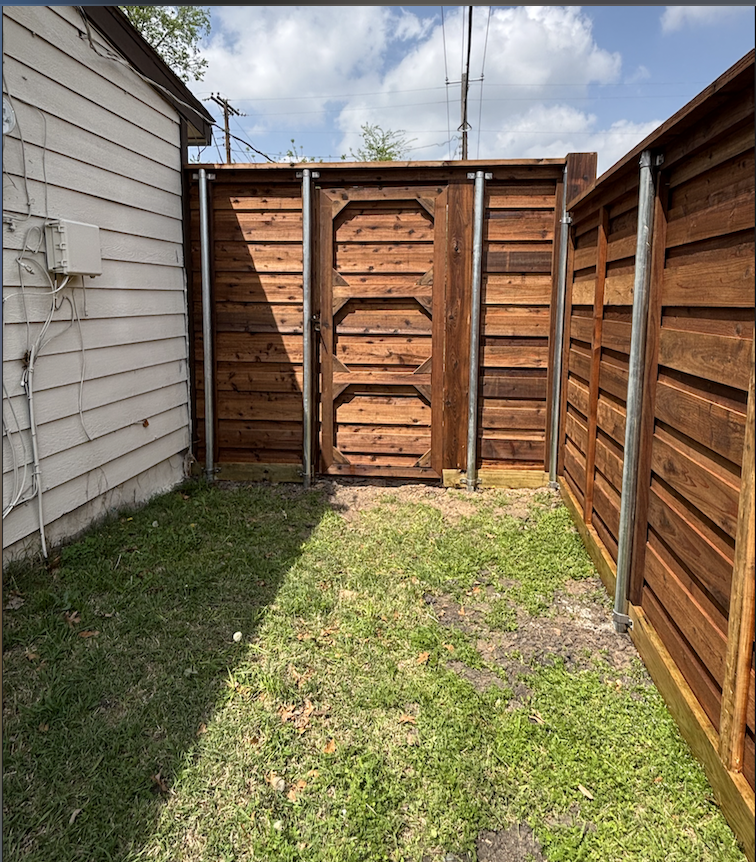Small backyard enclosed by wooden fence with a gate, with grass, some patches of dirt, and a white house side on the left with electrical wires.
