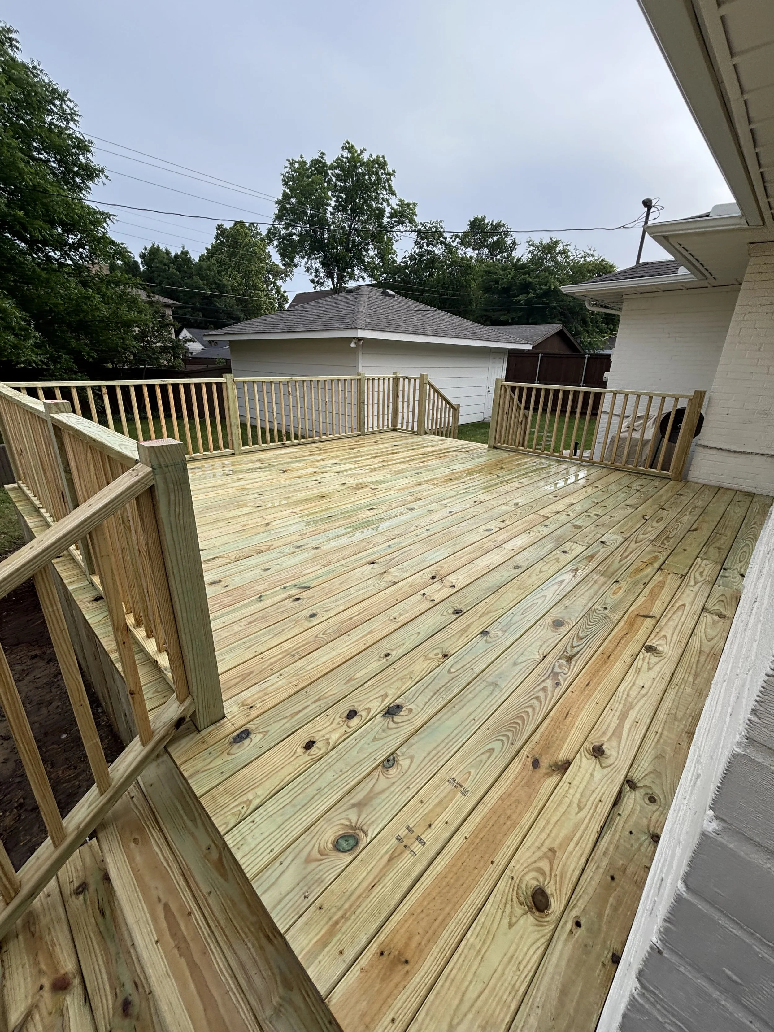 Freshly built wooden deck with new railing attached to a house, surrounded by backyard trees and neighboring structures.