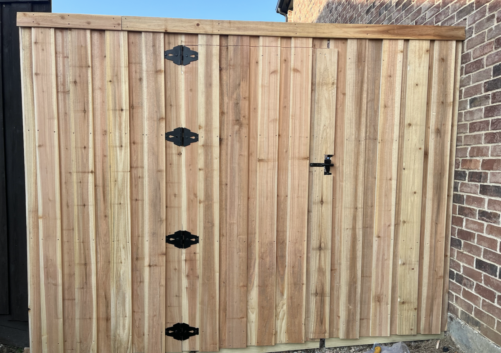 Freshly built wooden privacy fence with black hinges and latch, adjacent to a brick wall.