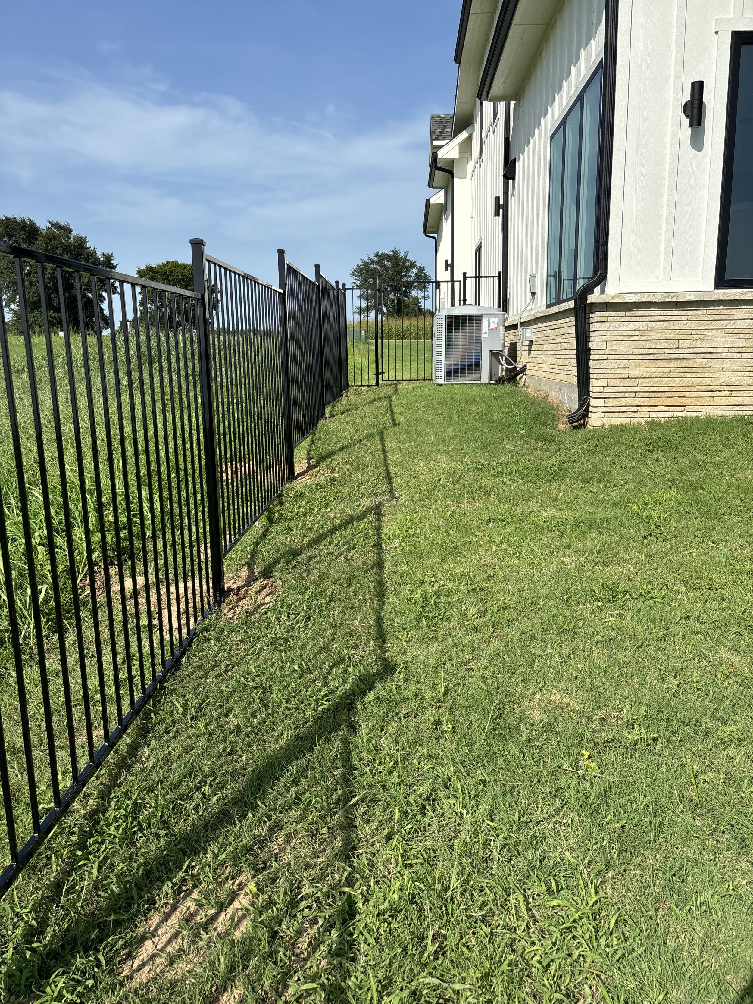 Side yard of a house with black metal fence, green grass, and an air conditioning unit near the house wall under large windows.