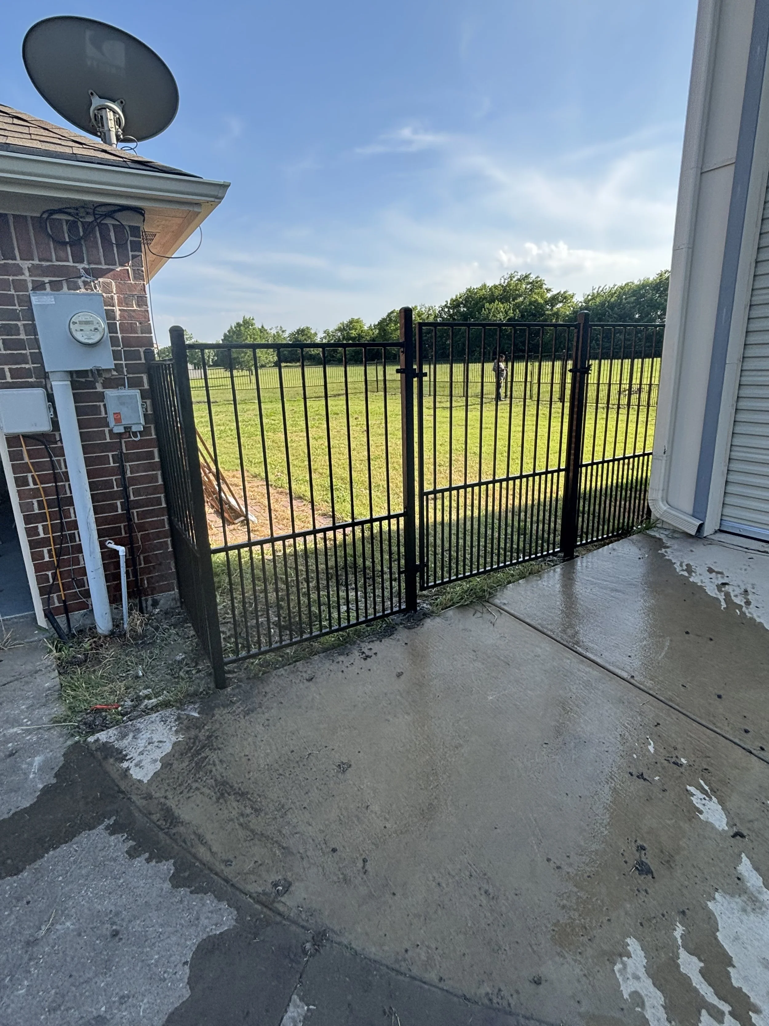 A black metal gate outside a house with a backyard and a person in the distance, blue sky with some clouds, wet concrete in the foreground, part of the house with a satellite dish and electrical meters.