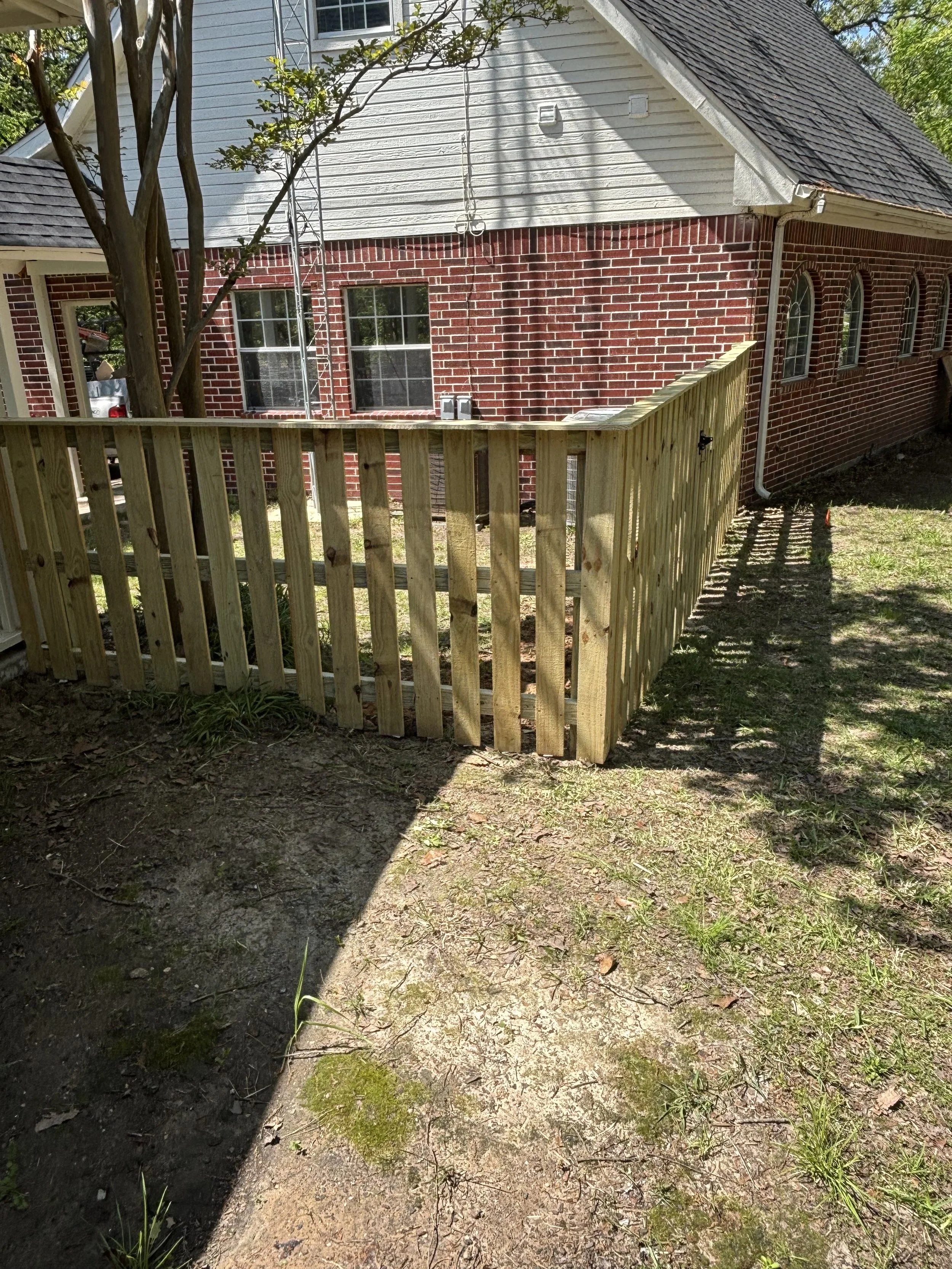 Newly built wooden fence in a backyard with brick house and trees in the background.