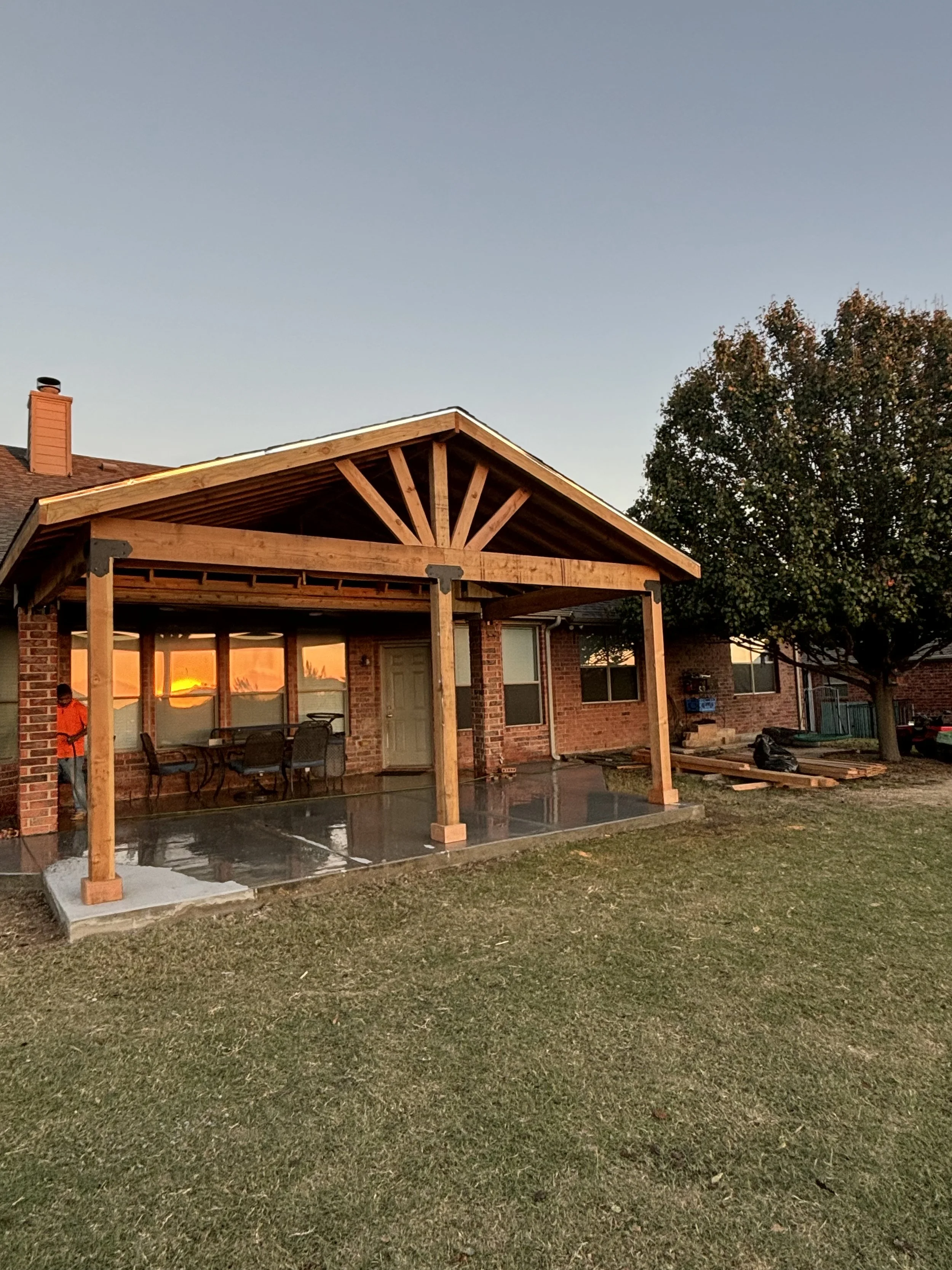 A house with a newly constructed wooden porch structure, featuring a sloped roof, supported by four wooden posts, with outdoor furniture and a chair visible underneath. A tree and other houses are seen in the background.