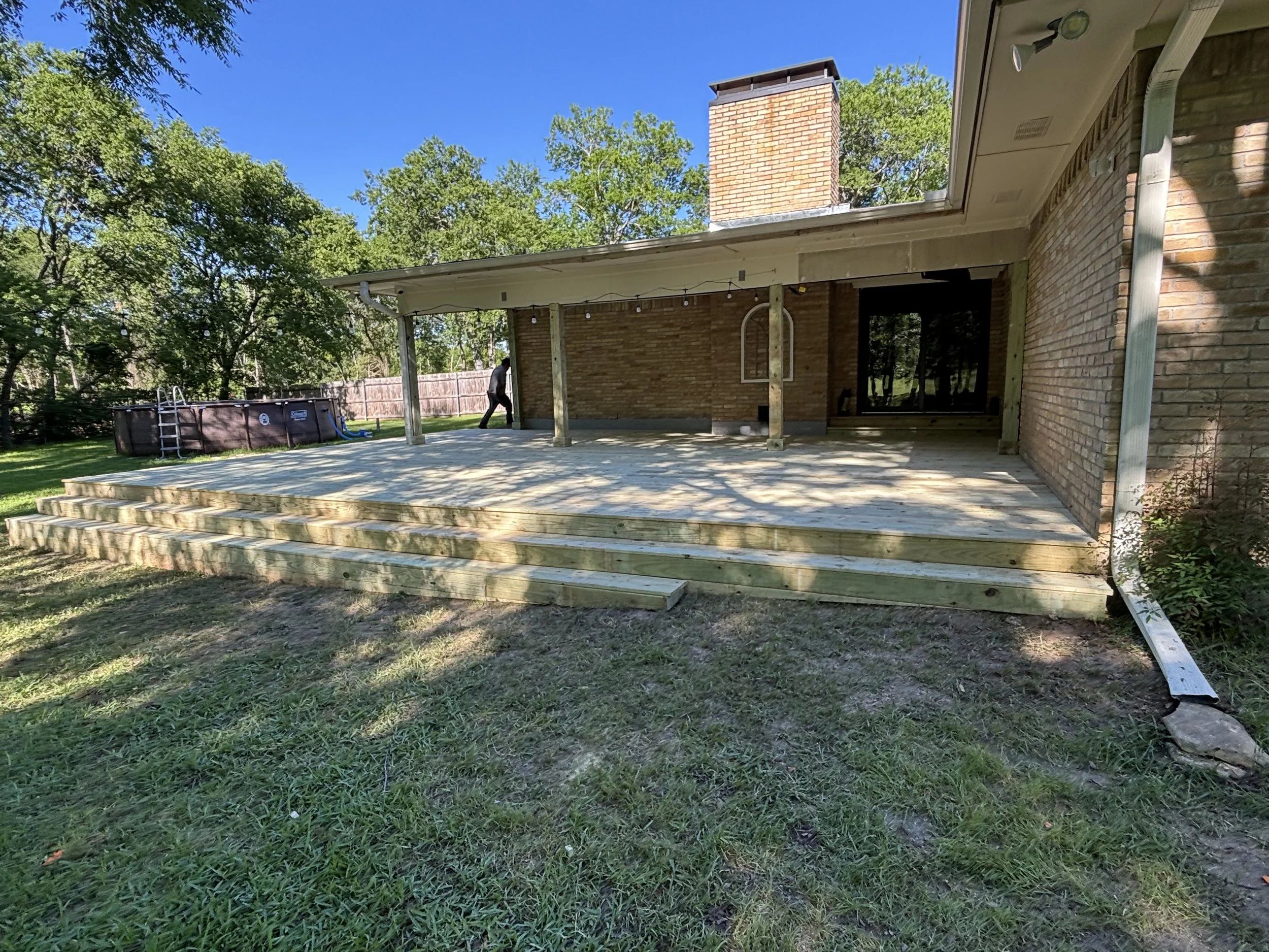 The image depicts a backyard with a newly built wooden deck attached to a brick house, with stairs leading down to the grass. A person is standing near the wood pile in the background, and a above ground pool is visible to the left. The sky is clear 