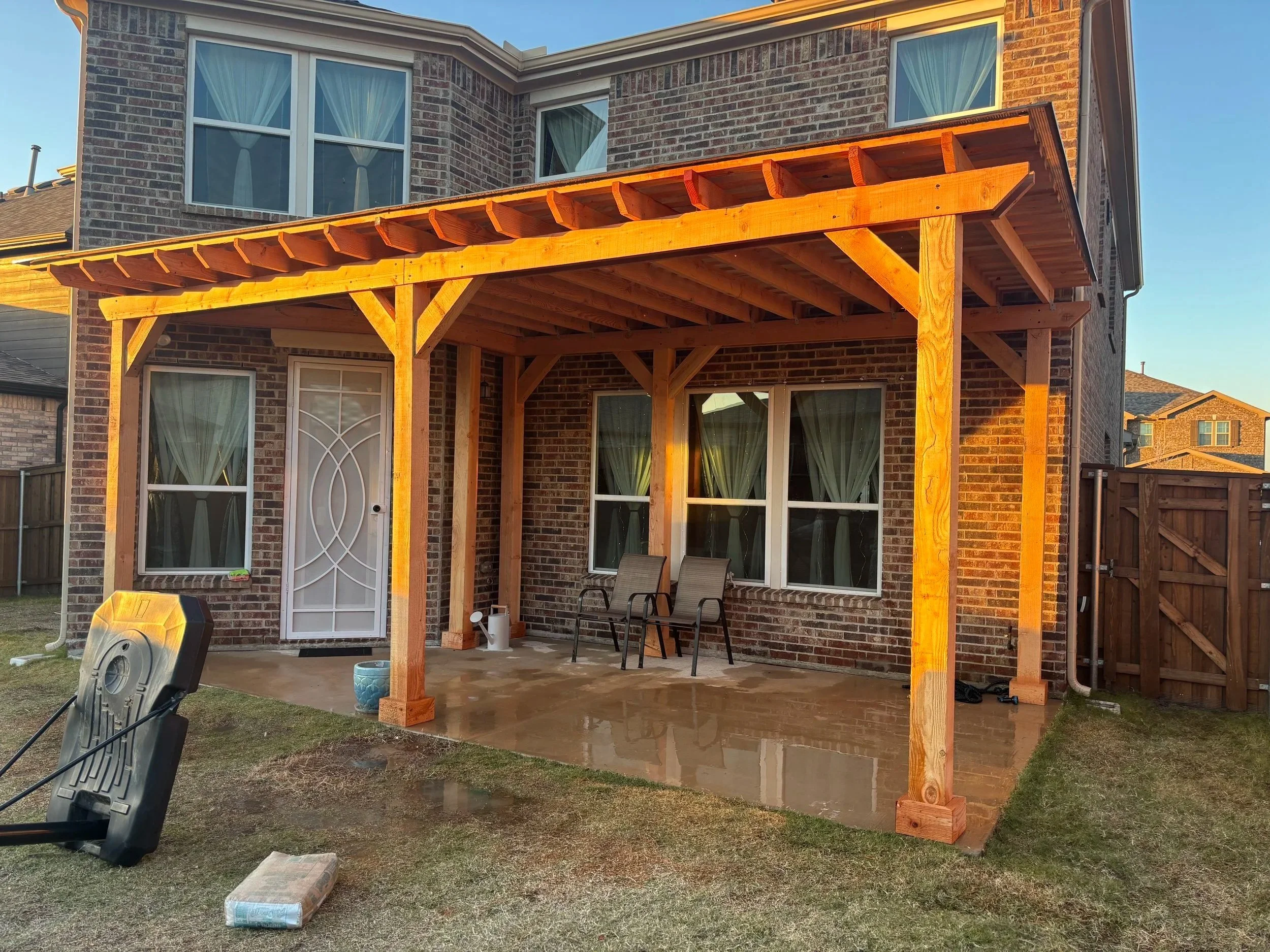 Backyard patio with a new wooden pergola attached to a brick house, two chairs under the pergola, and gardening tools nearby.