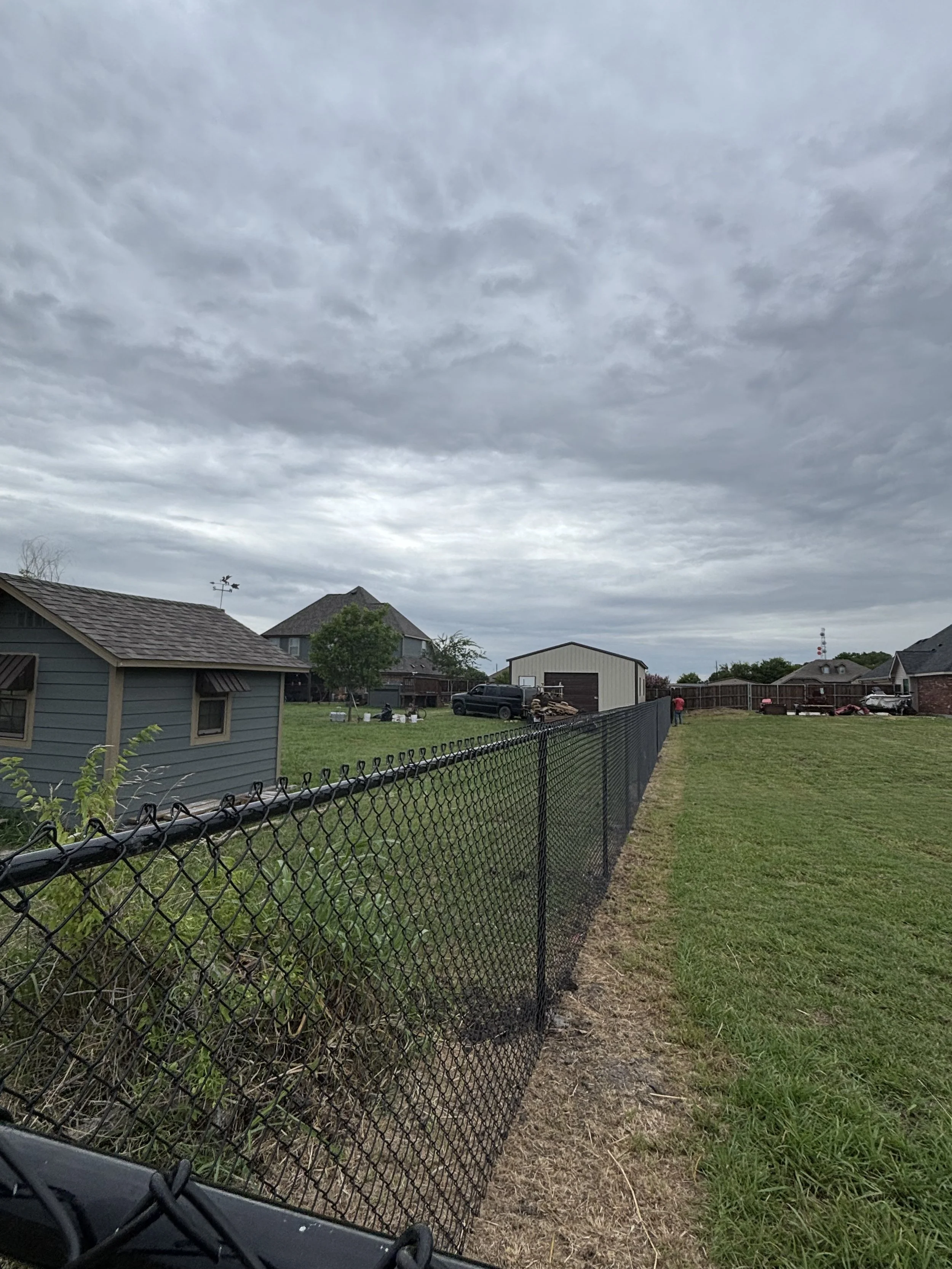 View of a backyard with a black chain-link fence, grass, and neighboring houses under an overcast sky.