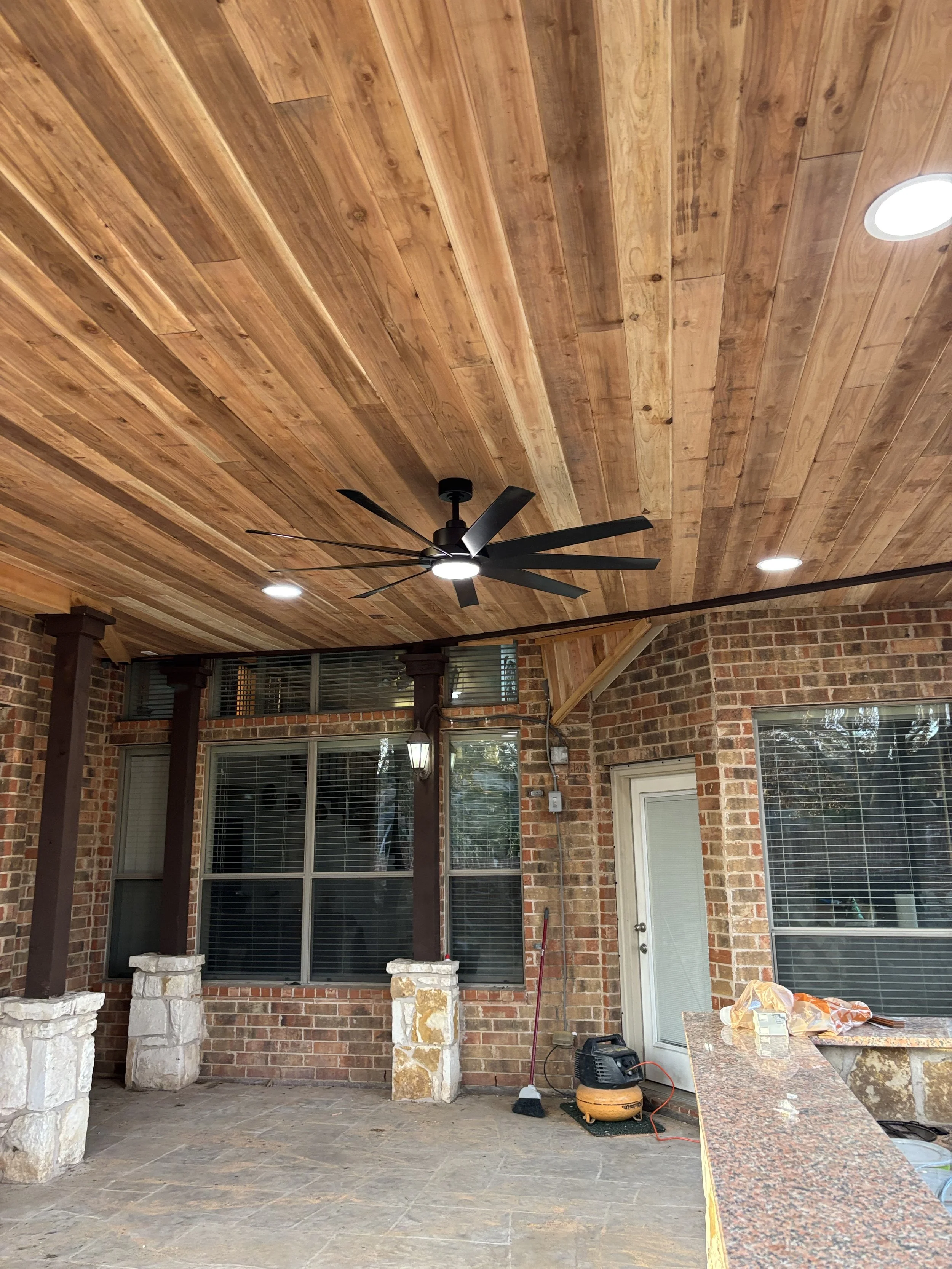 Photo of a covered patio area with a wooden ceiling, brick walls, large windows, and a ceiling fan with dark blades. The patio has construction tools, a broom, and a portable air compressor.