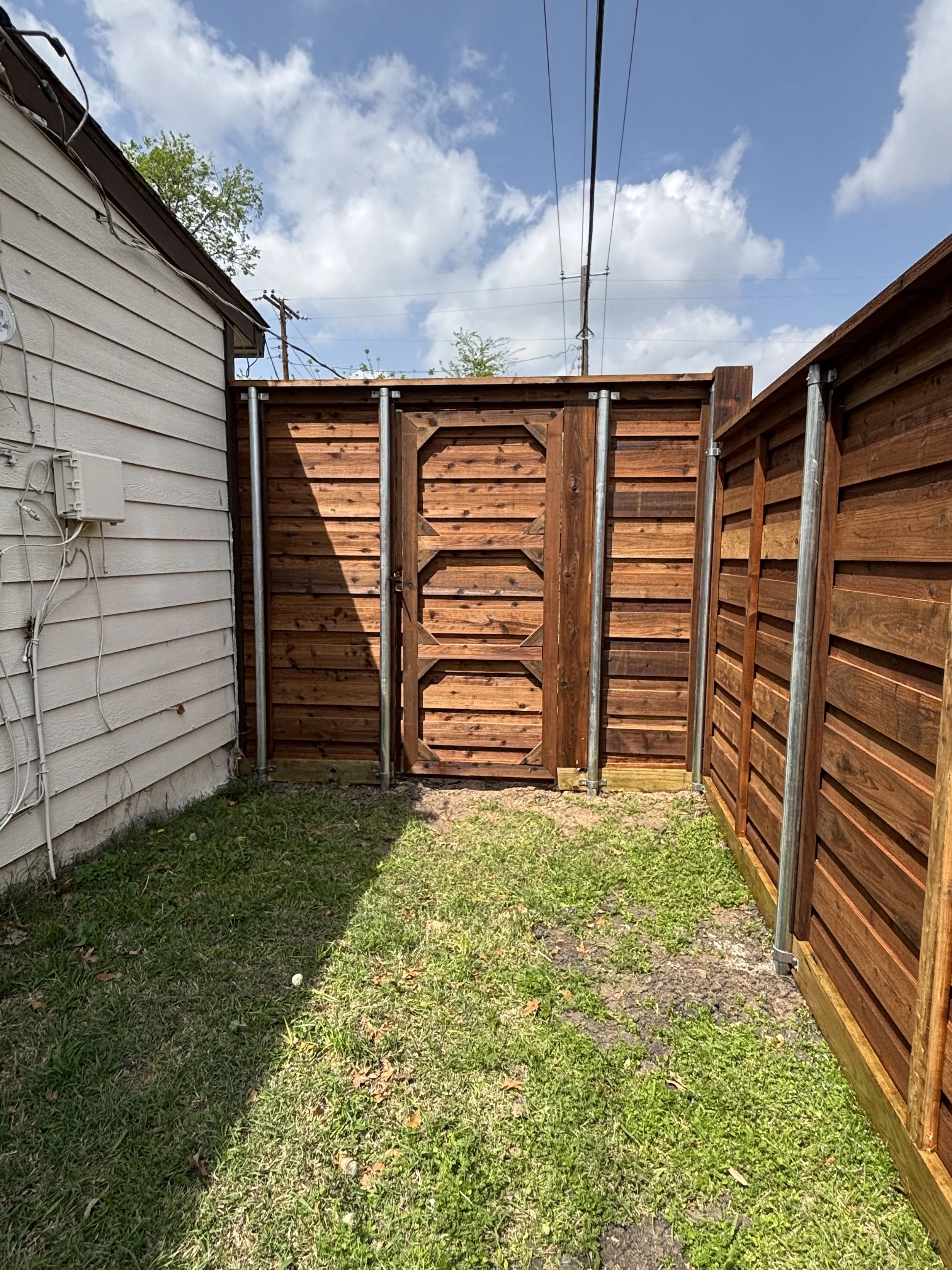 Backyard with a new wooden fence and gate, patches of grass and dirt, neighboring house on the left, blue sky with clouds, and utility wires overhead.