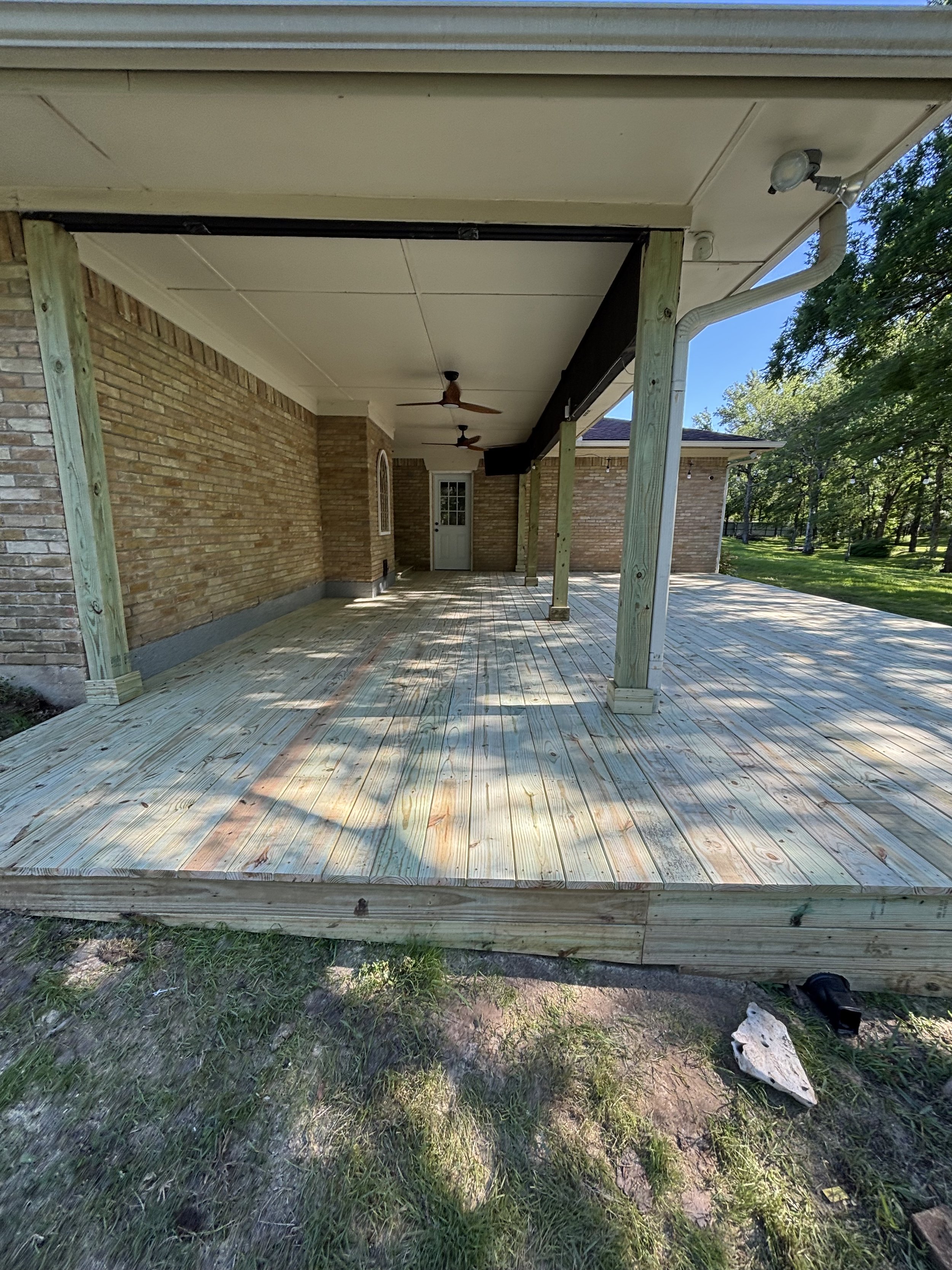 Newly constructed wooden deck attached to a brick house with ceiling fans, outdoor lighting, support beams, and surrounding green lawn and trees.