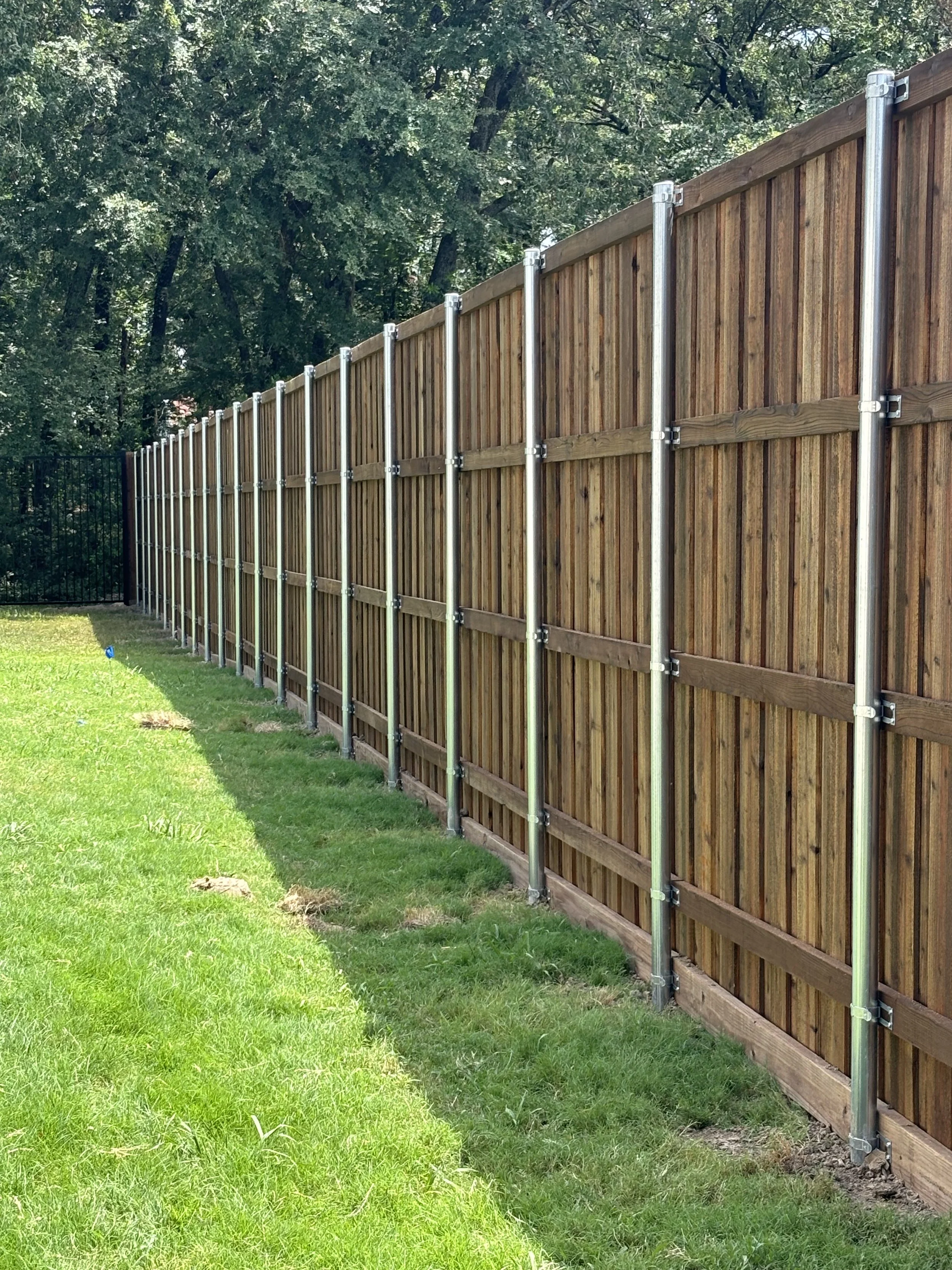 A newly installed wooden fence with metal posts along a grassy yard, with trees in the background.