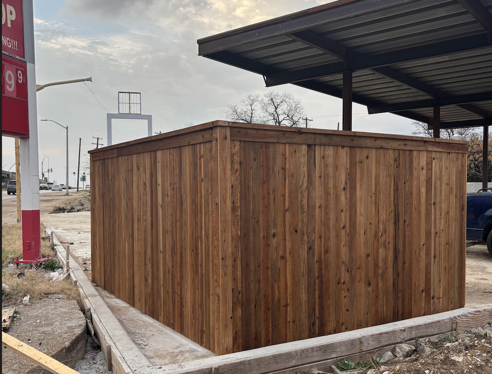A newly built wooden structure with vertical planks, situated on a concrete foundation, in an outdoor area near a parking lot and a canopy.