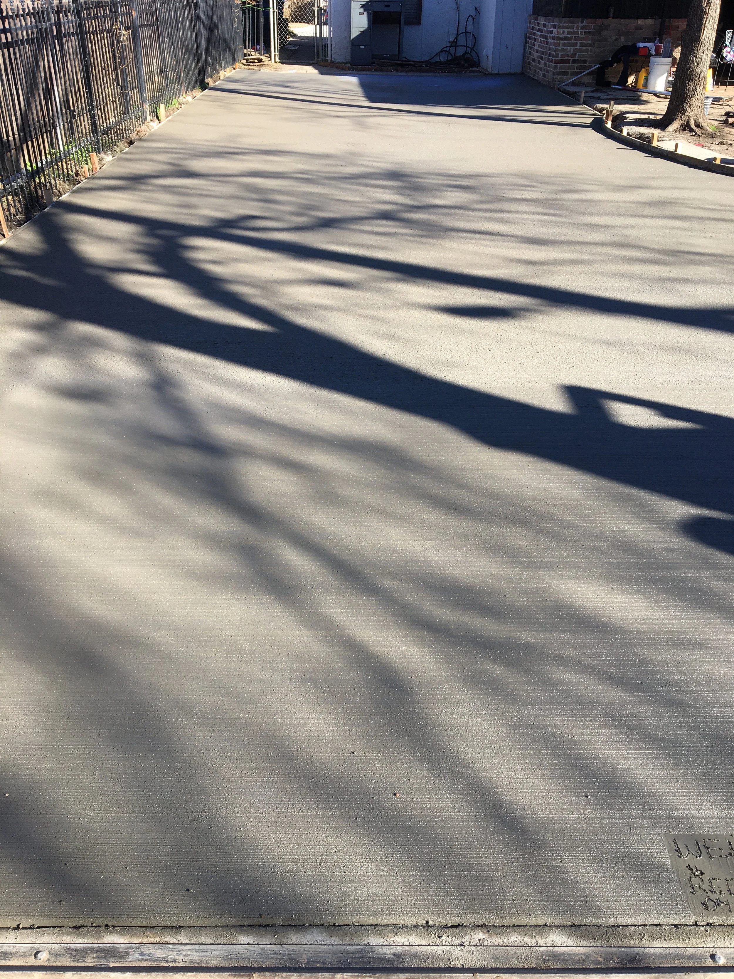 Freshly poured concrete slab in an outdoor backyard, with shadows from nearby trees cast across the surface, and a small section of handwritten note on the corner reading 'USE RE'.