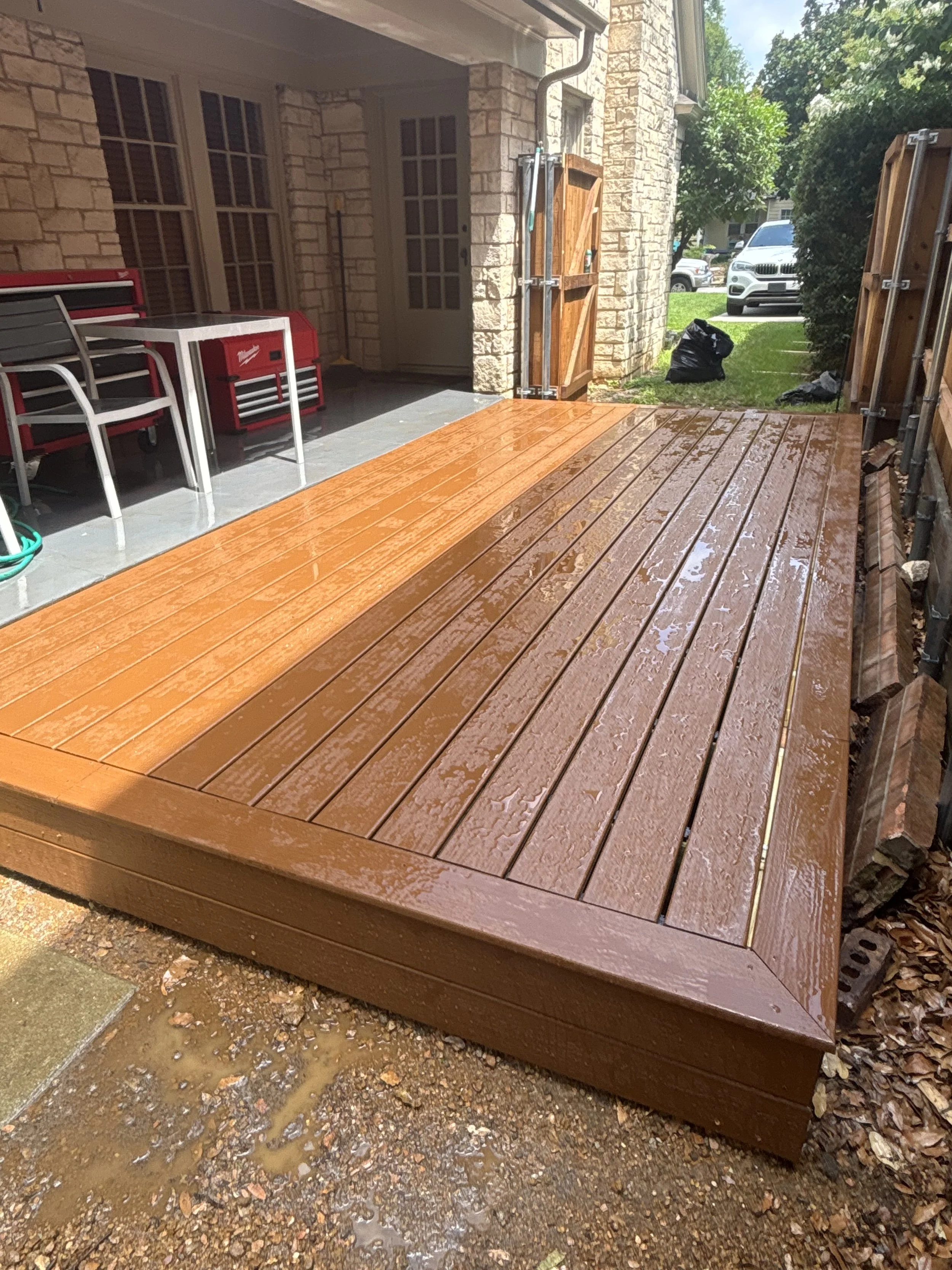 Newly installed wet wooden deck outside a house, with a brick wall, outdoor furniture, and a garden with trees and bushes in the background.