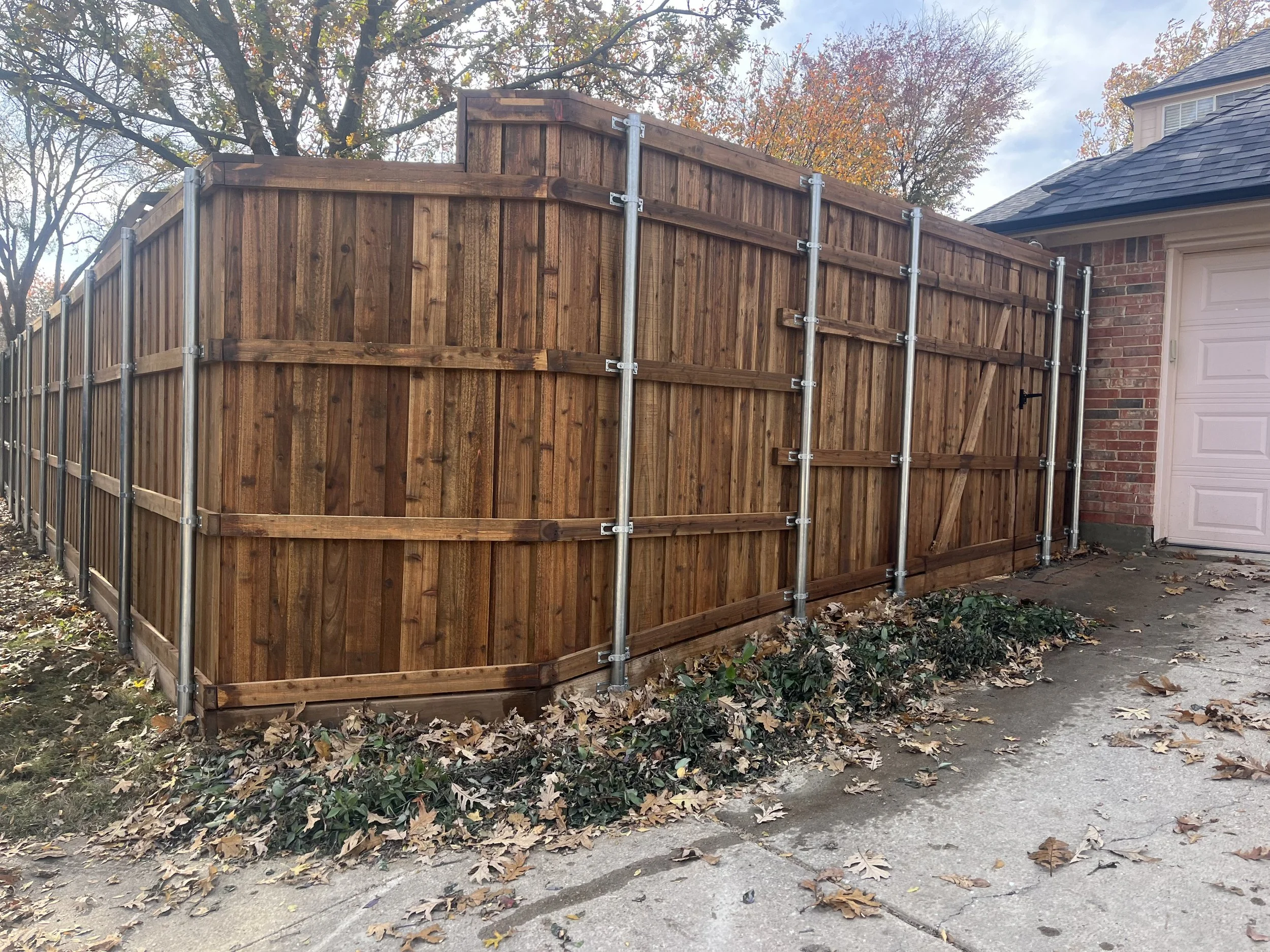 Newly installed wooden privacy fence with metal posts surrounds a driveway next to a brick house with a white garage door. Fallen leaves and ivy are on the ground.