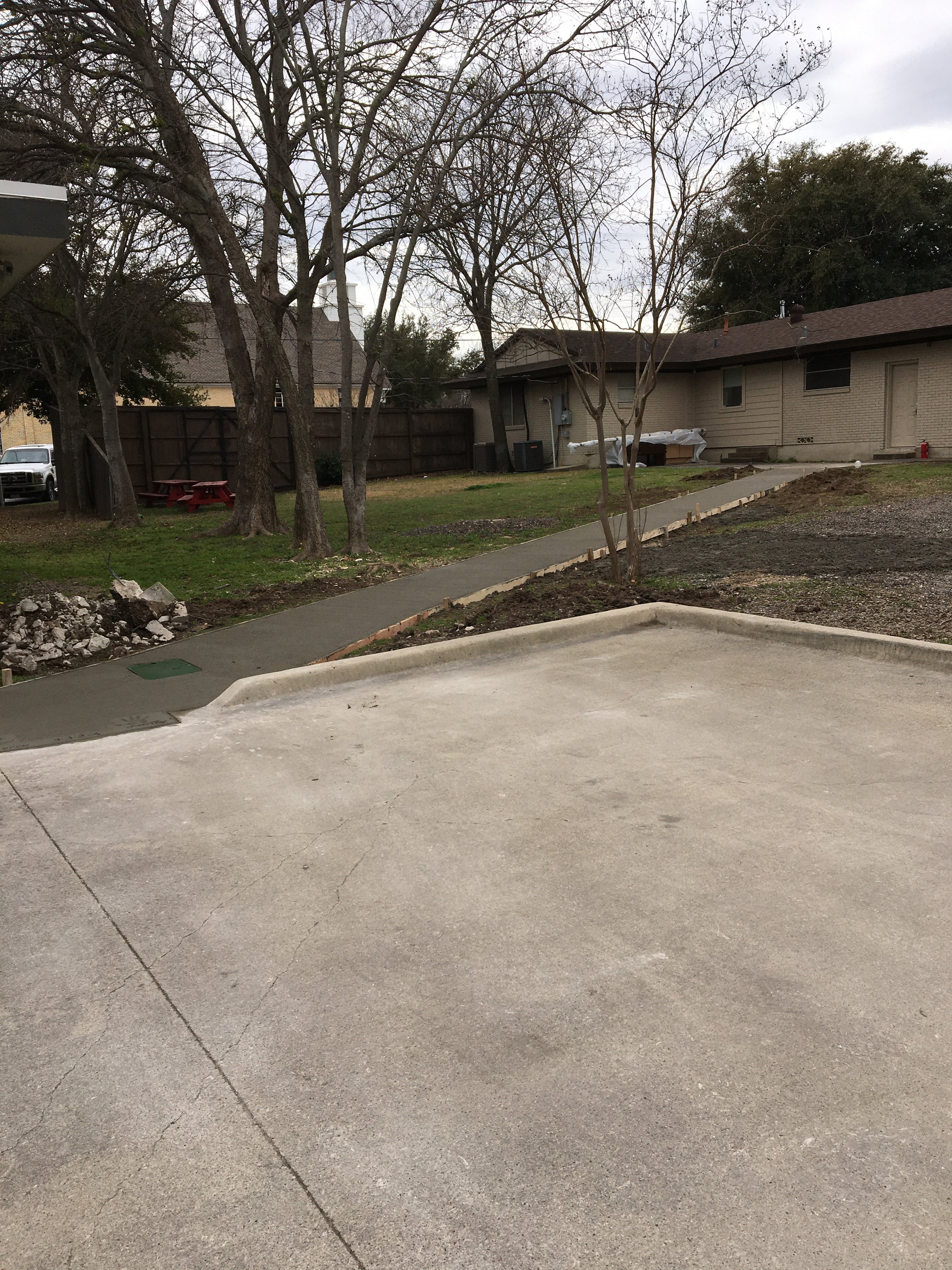 A backyard with a concrete sidewalk and pathway leading to a grassy area with leafless trees, a wooden fence, a covered outdoor area, and an unpaved section of dirt and gravel.