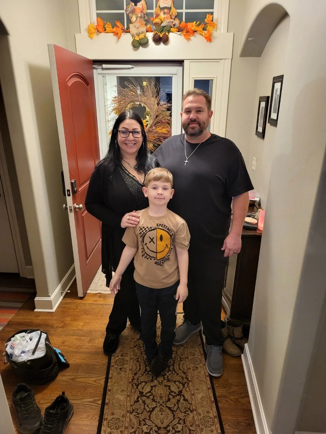 A family of three standing inside a home near the front door. The woman has long dark hair and glasses, wearing a black jacket. The man has short hair and a beard, wearing a black t-shirt with a cross necklace. The young boy in front has short blonde hair, wearing a beige T-shirt with a smiley face design and black pants. The door is open, showing a fall-themed wreath outside, and there are autumn decorations on the window ledge above with stuffed dolls and leaves. Shoes and a bag are on the floor nearby.