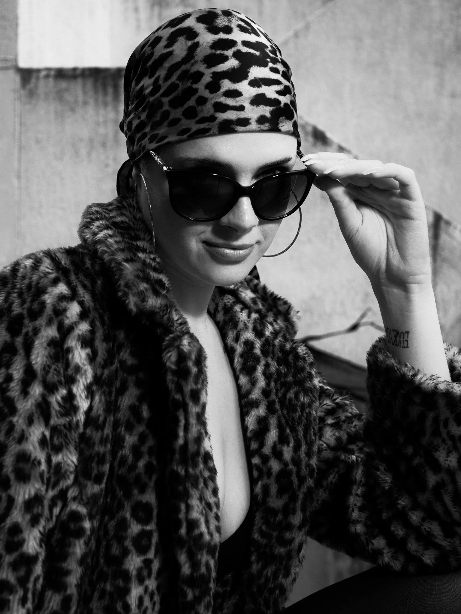 A woman wearing a leopard-print headscarf, sunglasses, and a leopard-print coat, with her hand adjusting her sunglasses.