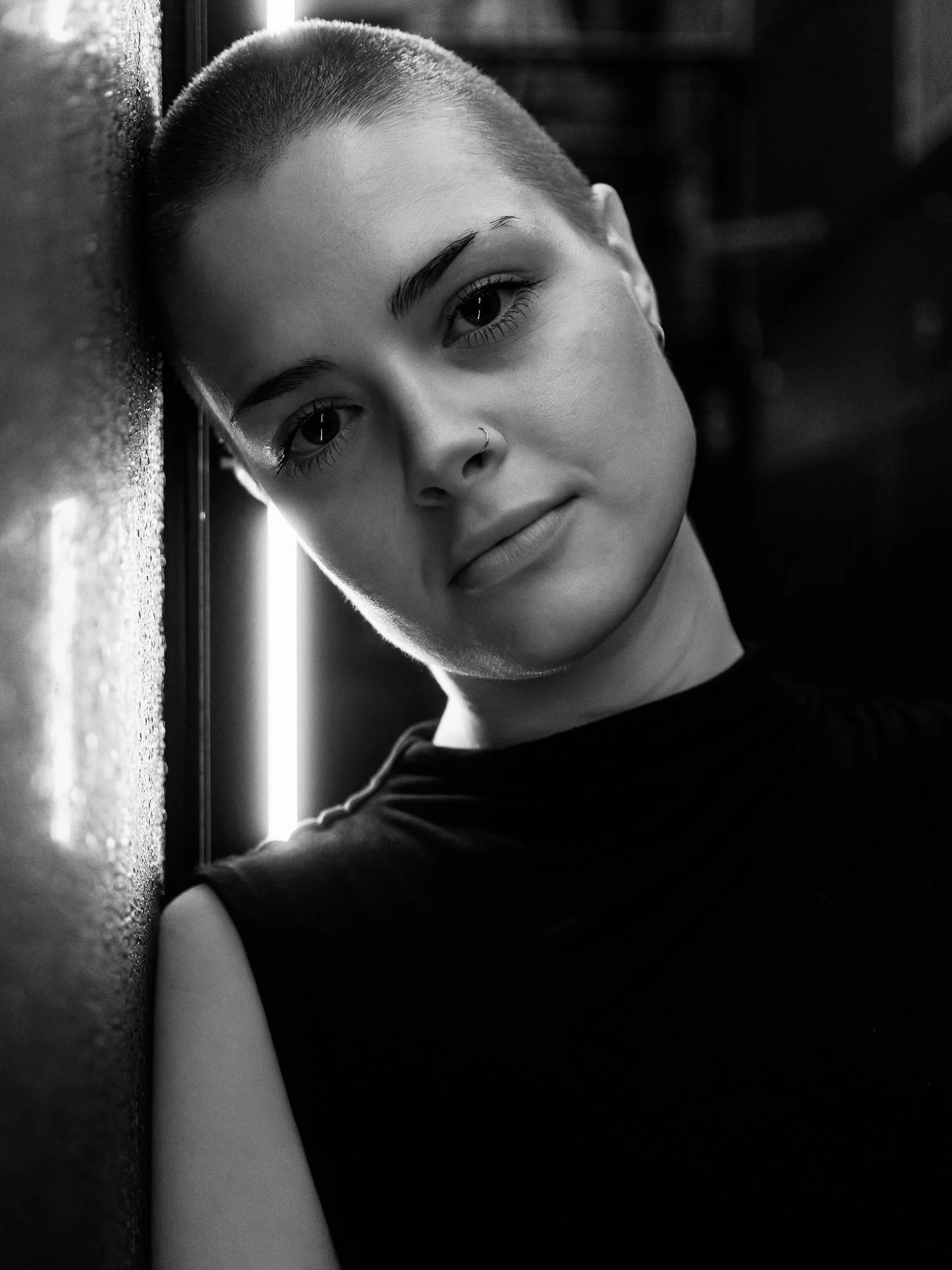 A young woman with a short buzz cut hairstyle looking directly at the camera, leaning against a textured wall with a vertical light source behind her. The photo is in black and white.