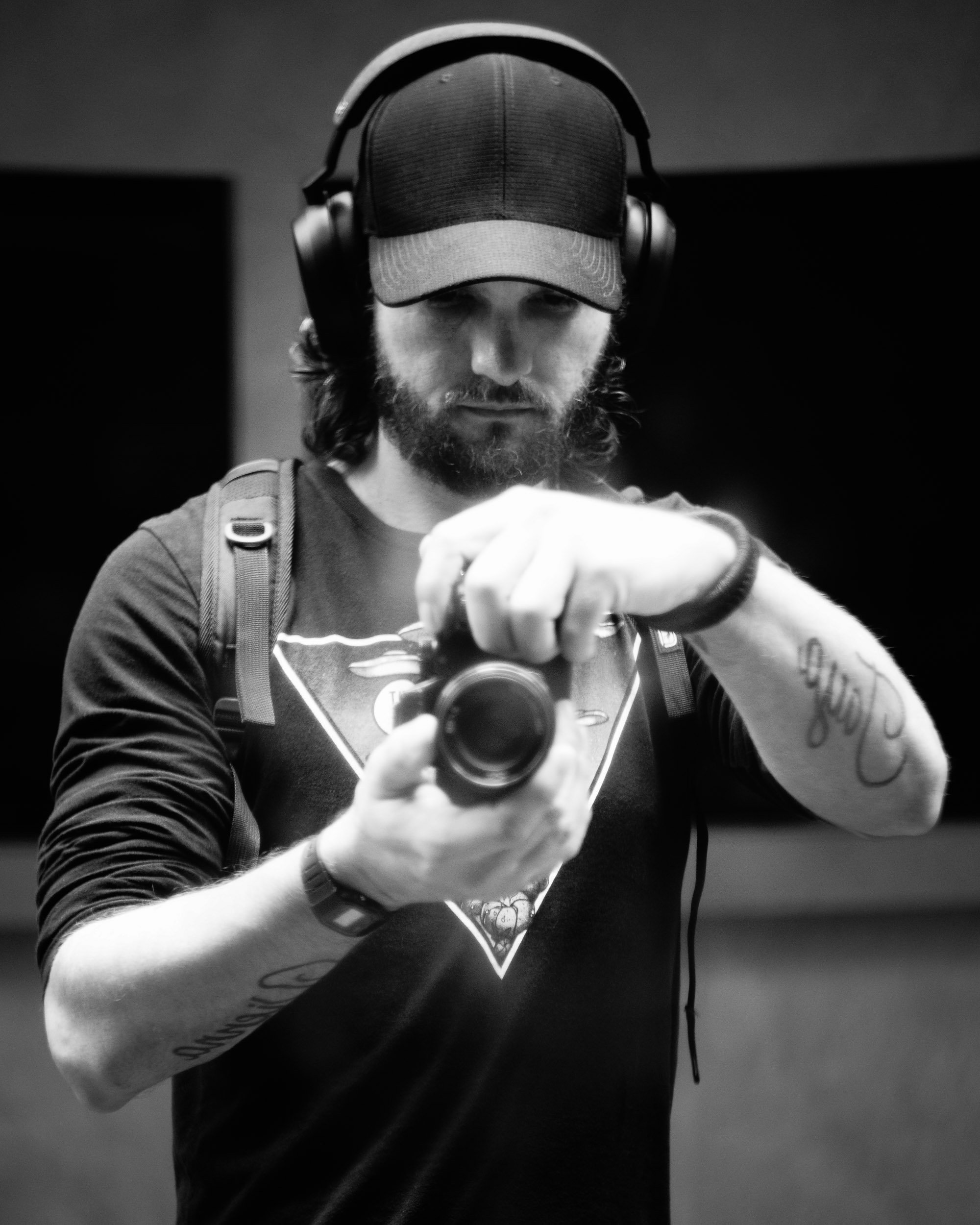 Black and white photo of a man wearing a cap and headphones, taking a selfie with a camera, with tattoos on his arms.