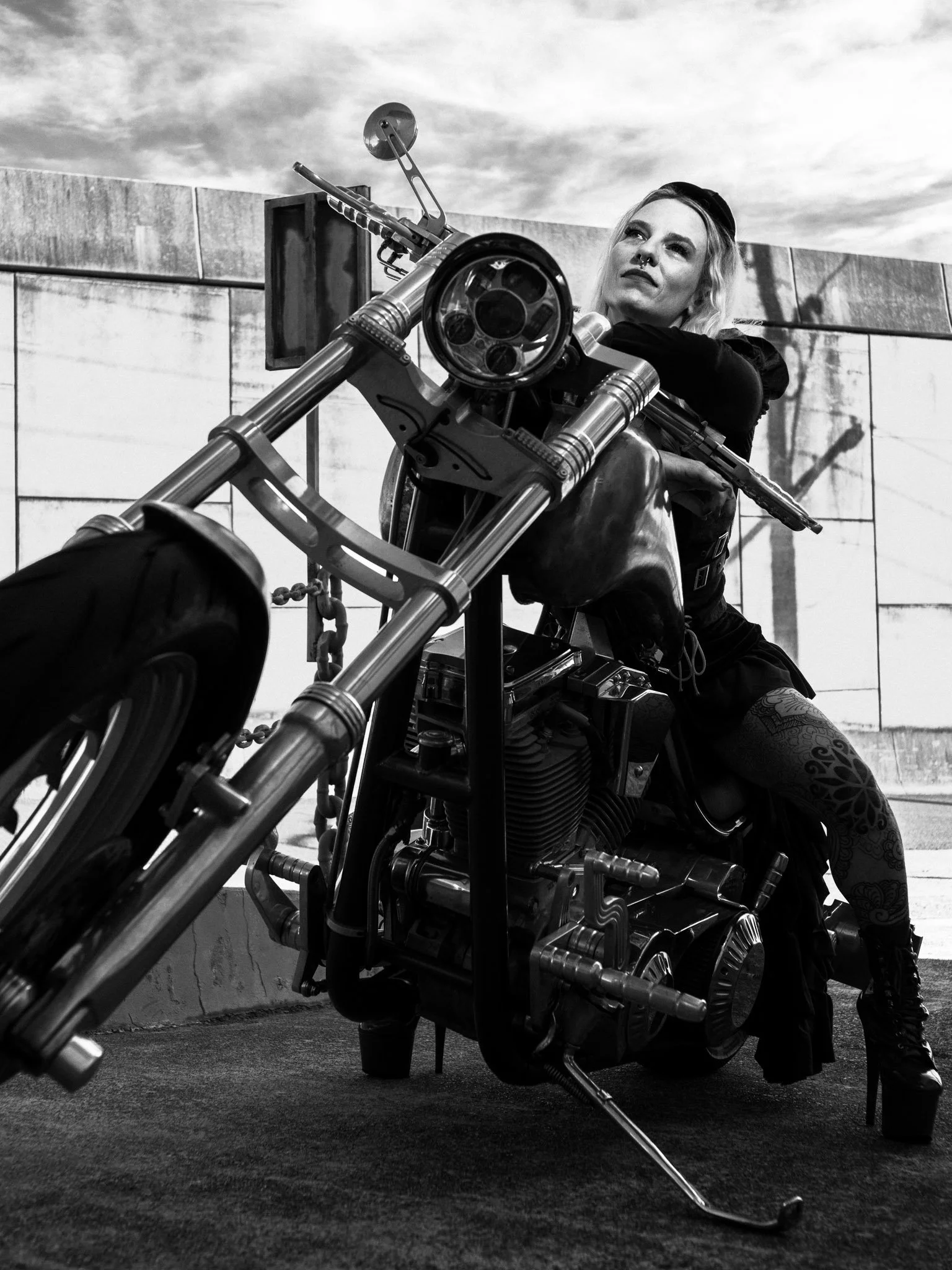 A woman with tattoos wearing high-heeled boots sitting on a custom motorcycle beneath a cloudy sky with a concrete wall in the background.