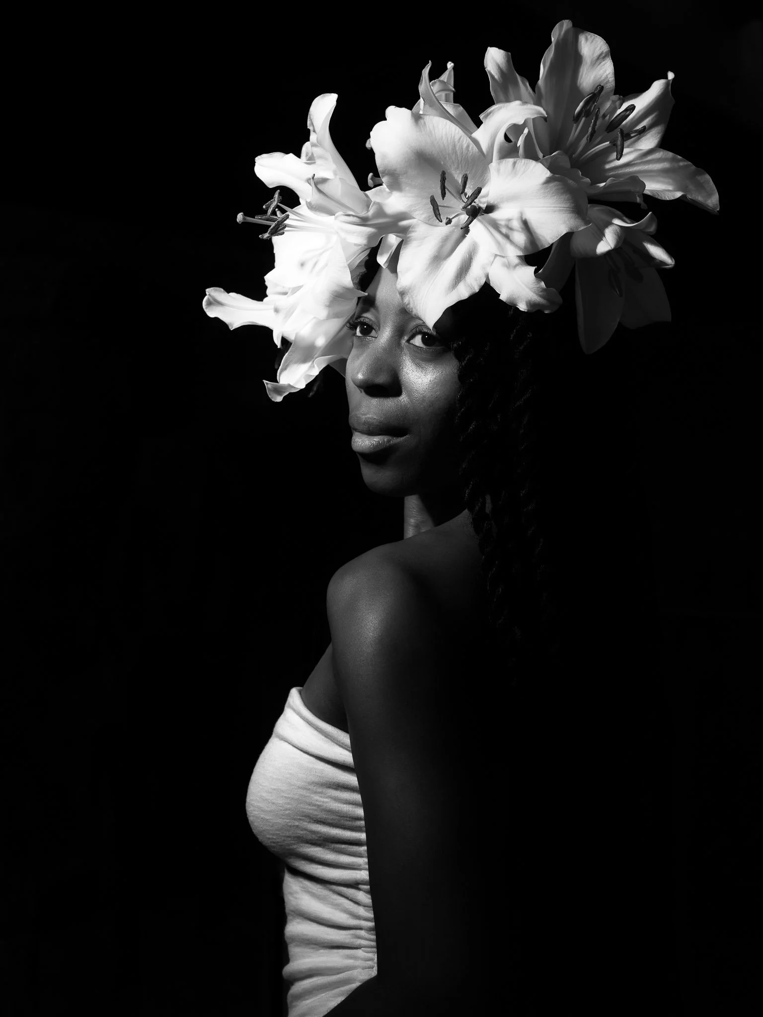 A black and white portrait of a woman with flowers on her head, looking to the side, with a dark background.