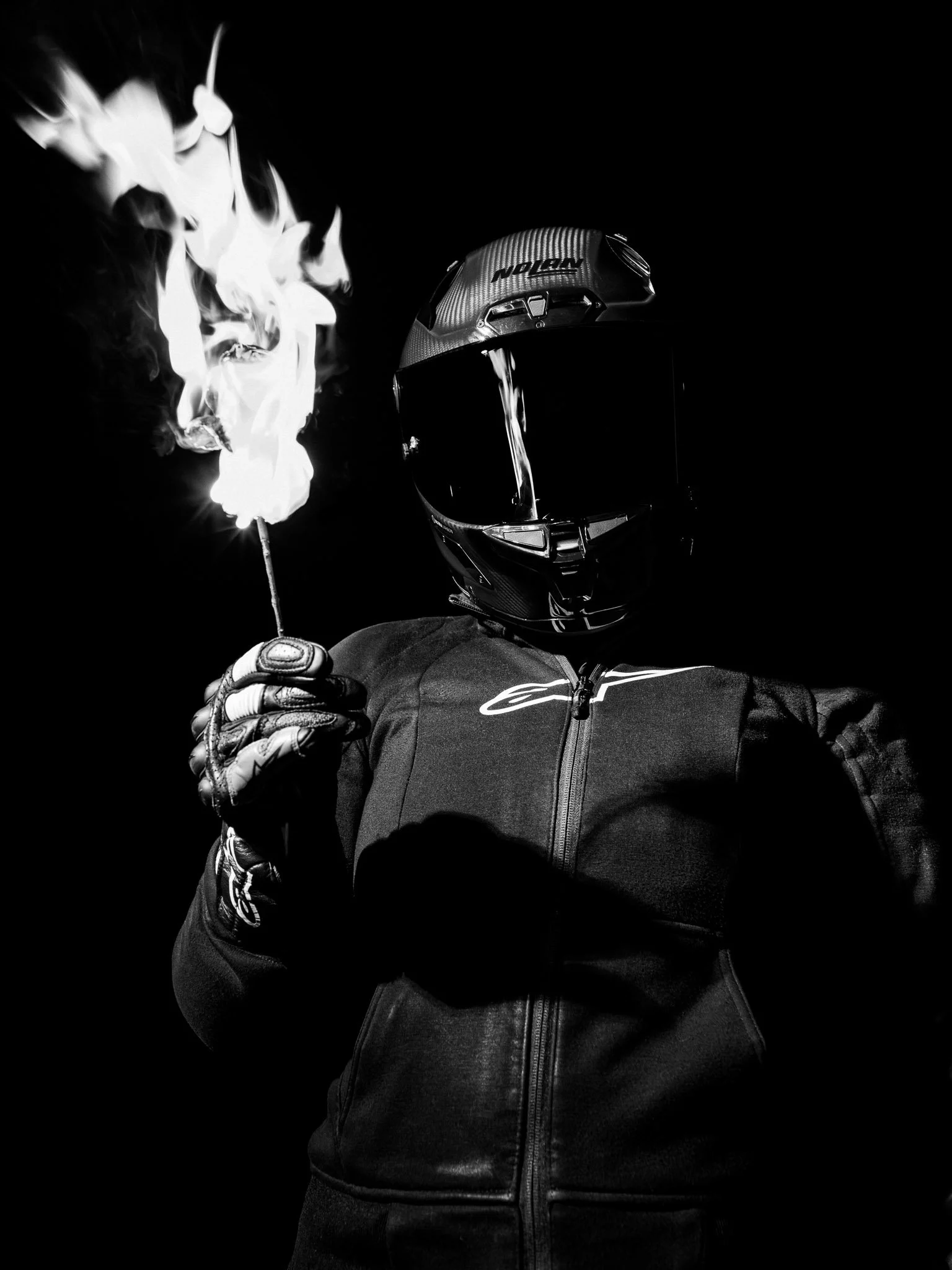 Person wearing a motorcycle helmet and gloves, holding a lit match with flame, in a dark setting.