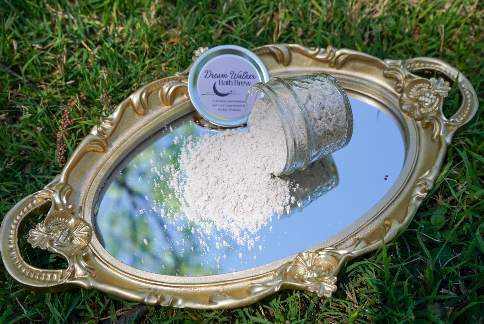 Handmade Dream Walker bath brew spilled from a glass jar onto a gold vintage mirror tray outdoors on grass.