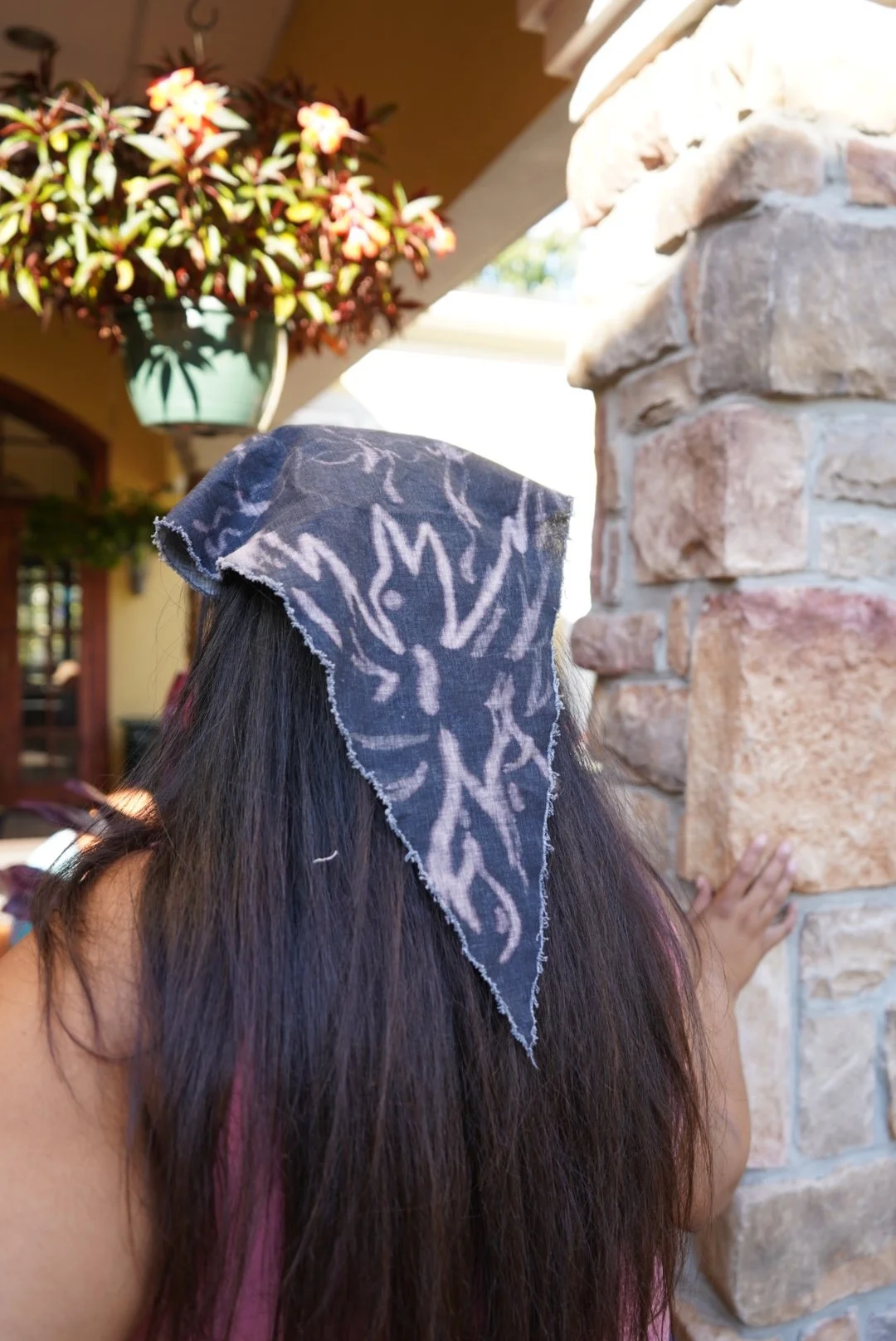 Bleach-painted dark headscarf with frayed edges worn outdoors near a stone wall with hanging flowers in sunlight.