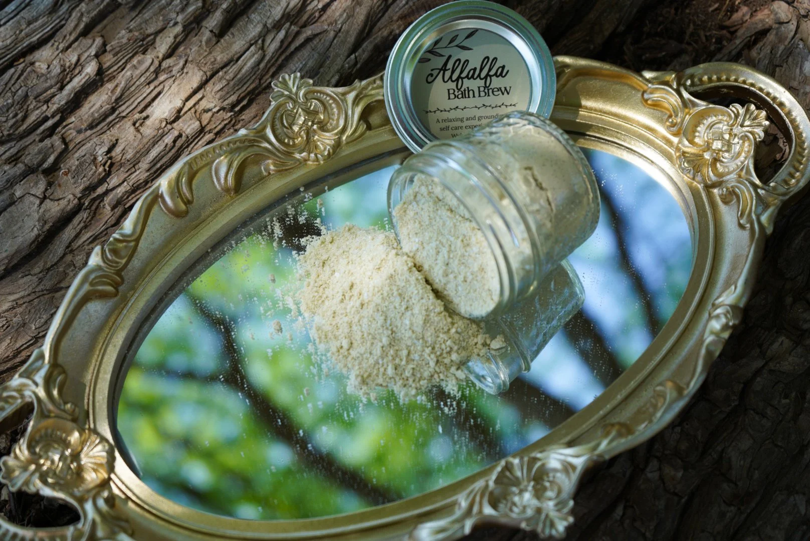 Alfalfa bath brew spilled from a glass jar onto a vintage gold mirror tray outdoors with natural greenery reflections.