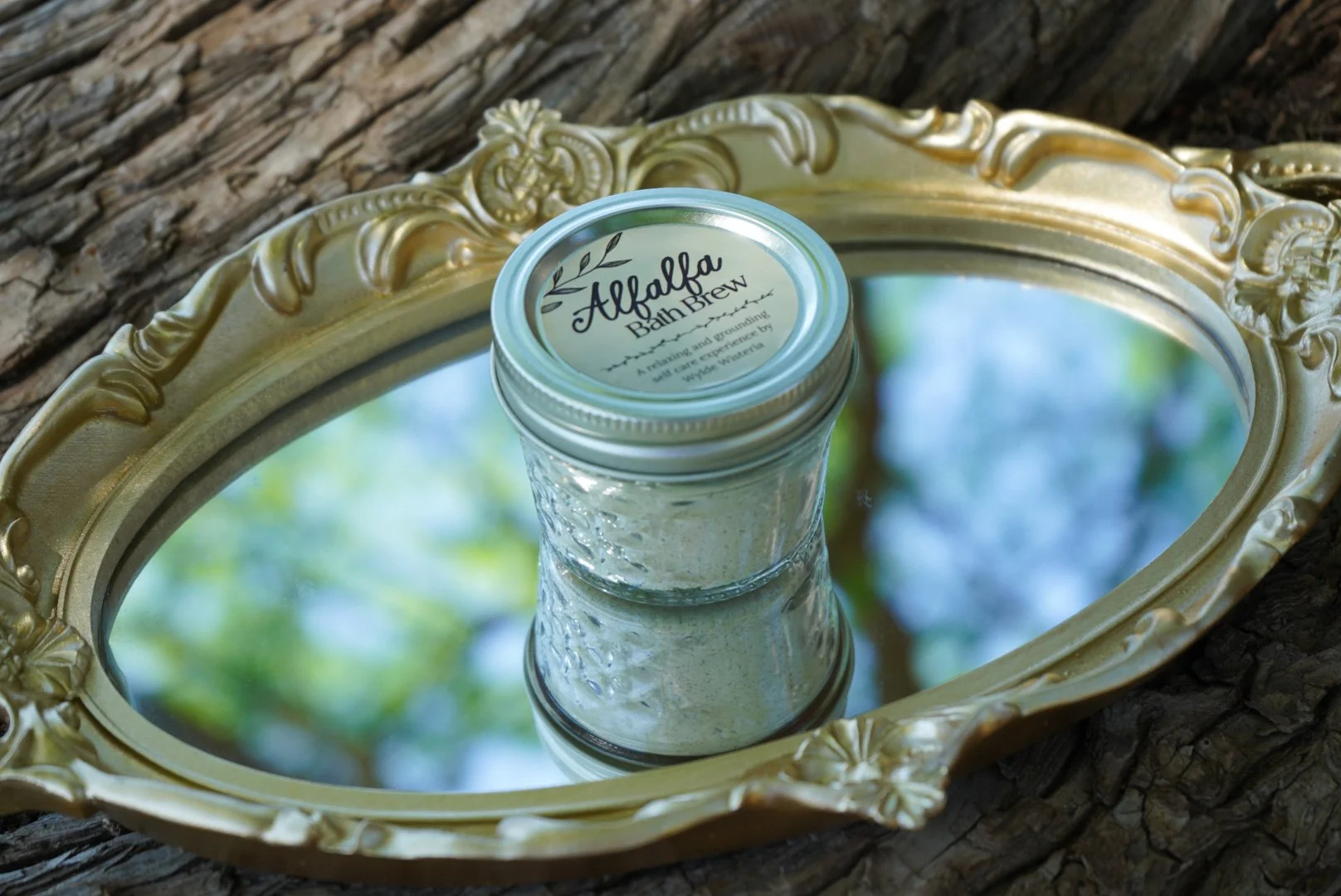 Alfalfa bath brew jar on a vintage gold mirror tray outdoors on wood with natural greenery reflections.