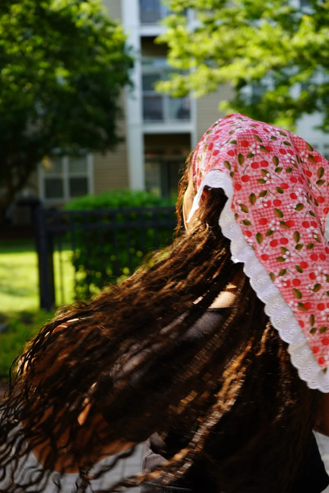 Red gingham cherry-print veil with white lace trim worn outdoors in sunlight with greenery in the background.