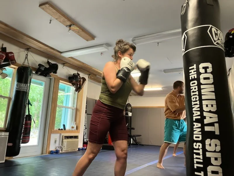 A woman practicing boxing in a gym, wearing gloves and in a fighting stance near a punching bag, with a man in the background also training.