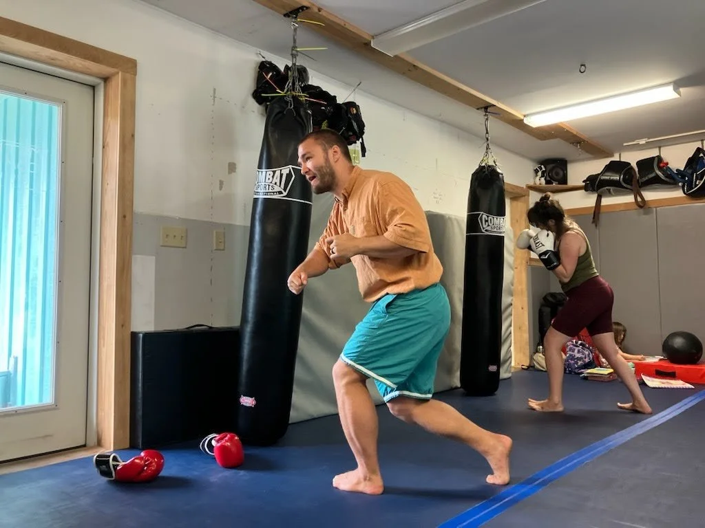 A man and a woman practicing boxing in a gym with hanging punching bags. The gym has black punching bags, helmets, and other training equipment.