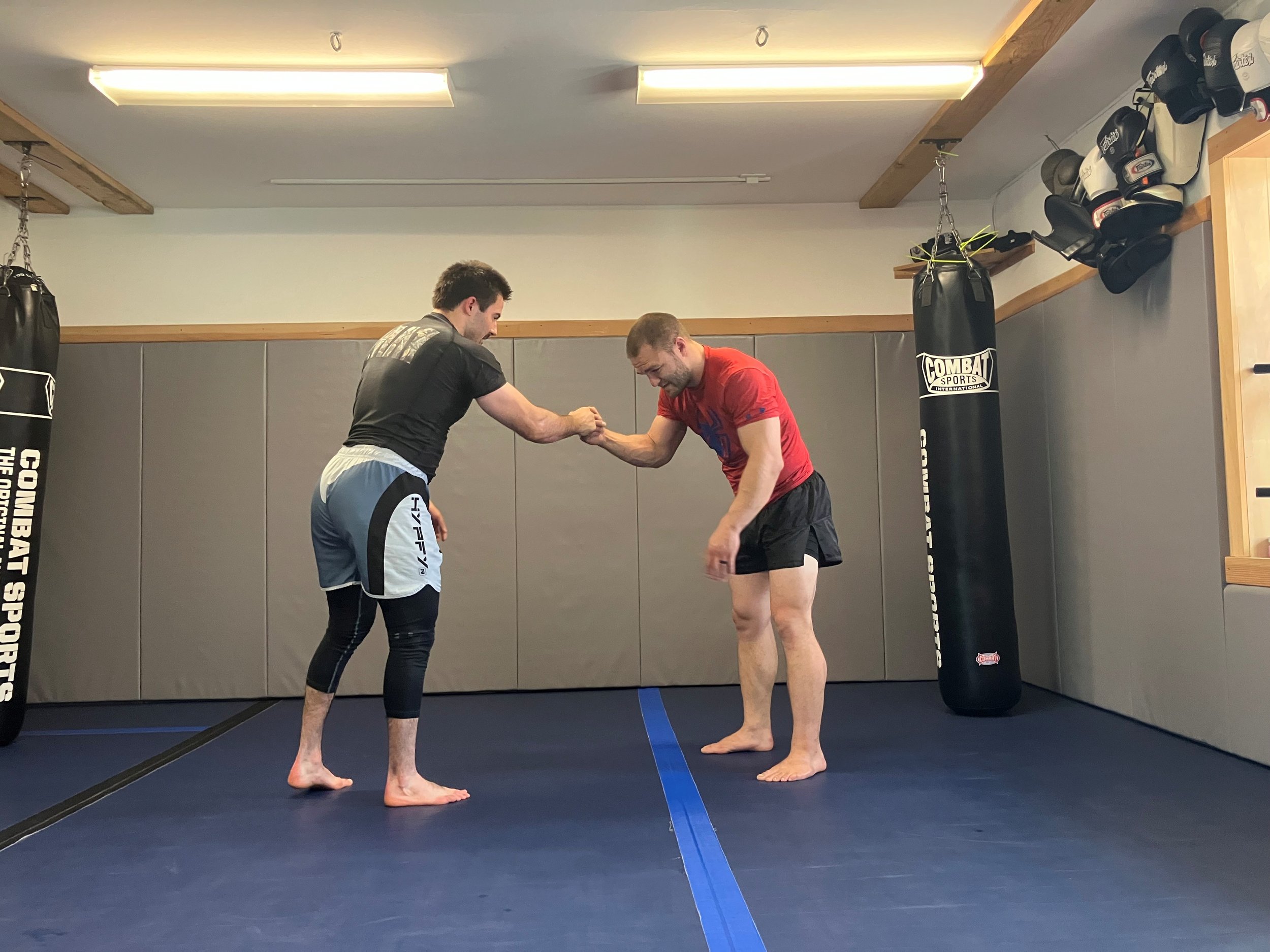 Two men practicing martial arts in a gym, engaging in a handshake or grip during training, with punching bags and boxing gloves on the wall.