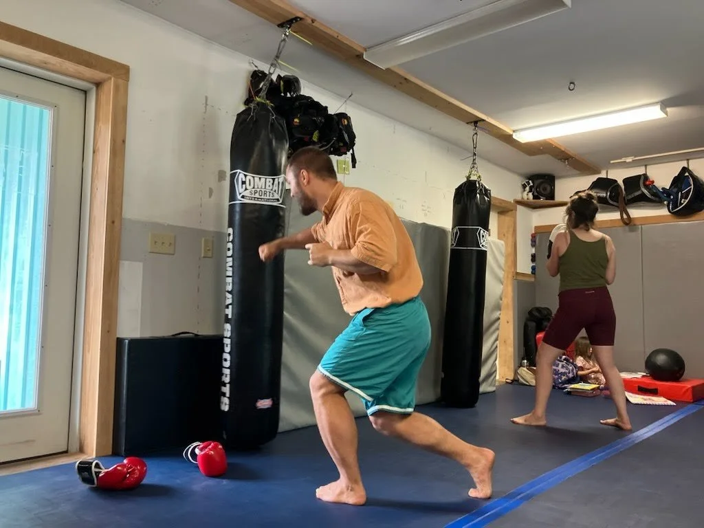 A man punches at a punching bag in a gym, while a woman exercises nearby. The gym has boxing equipment, including gloves and punching bags.